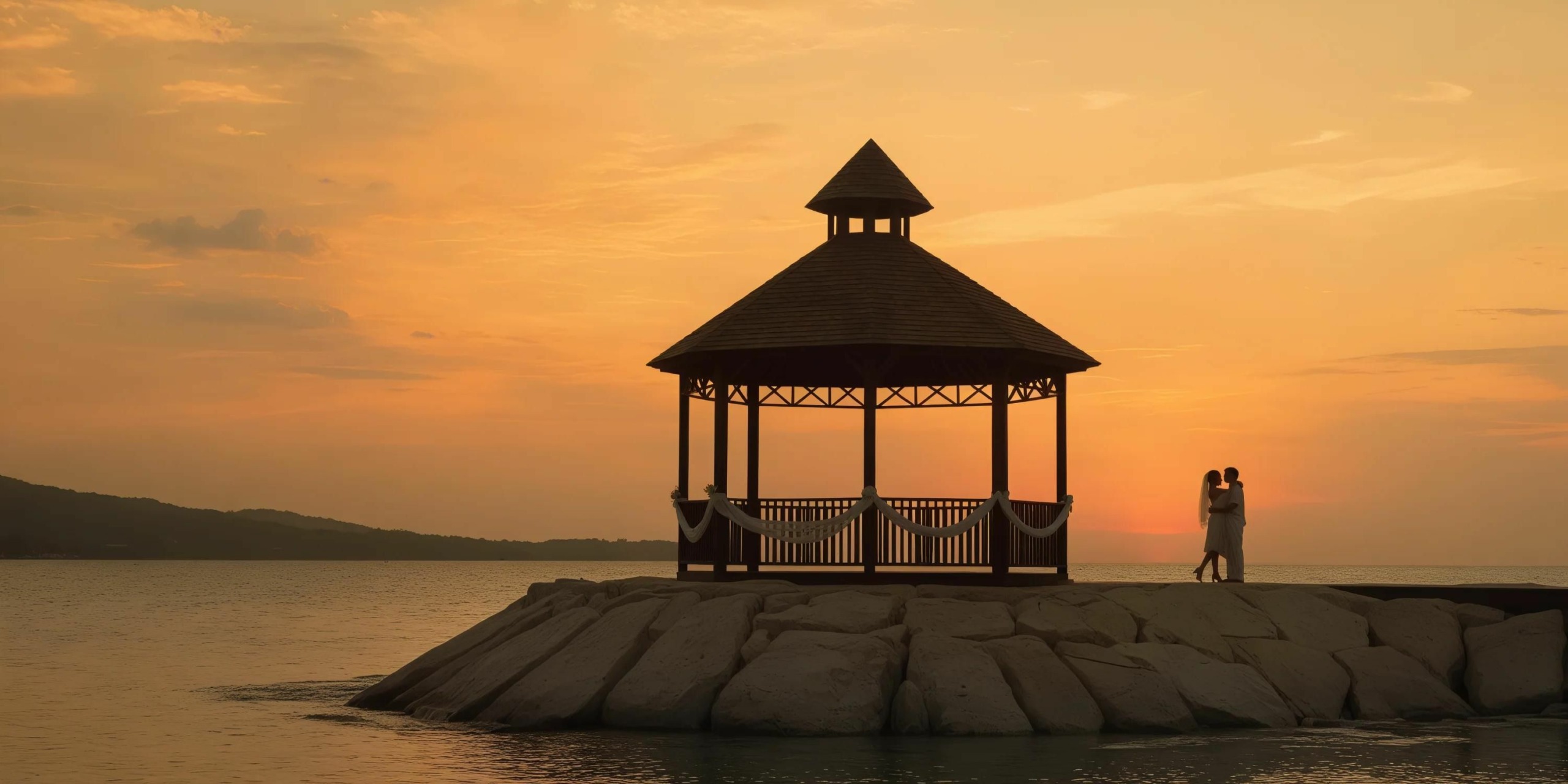 A couple stands embracing under a wooden gazebo at the edge of a rocky shoreline during a sunset.