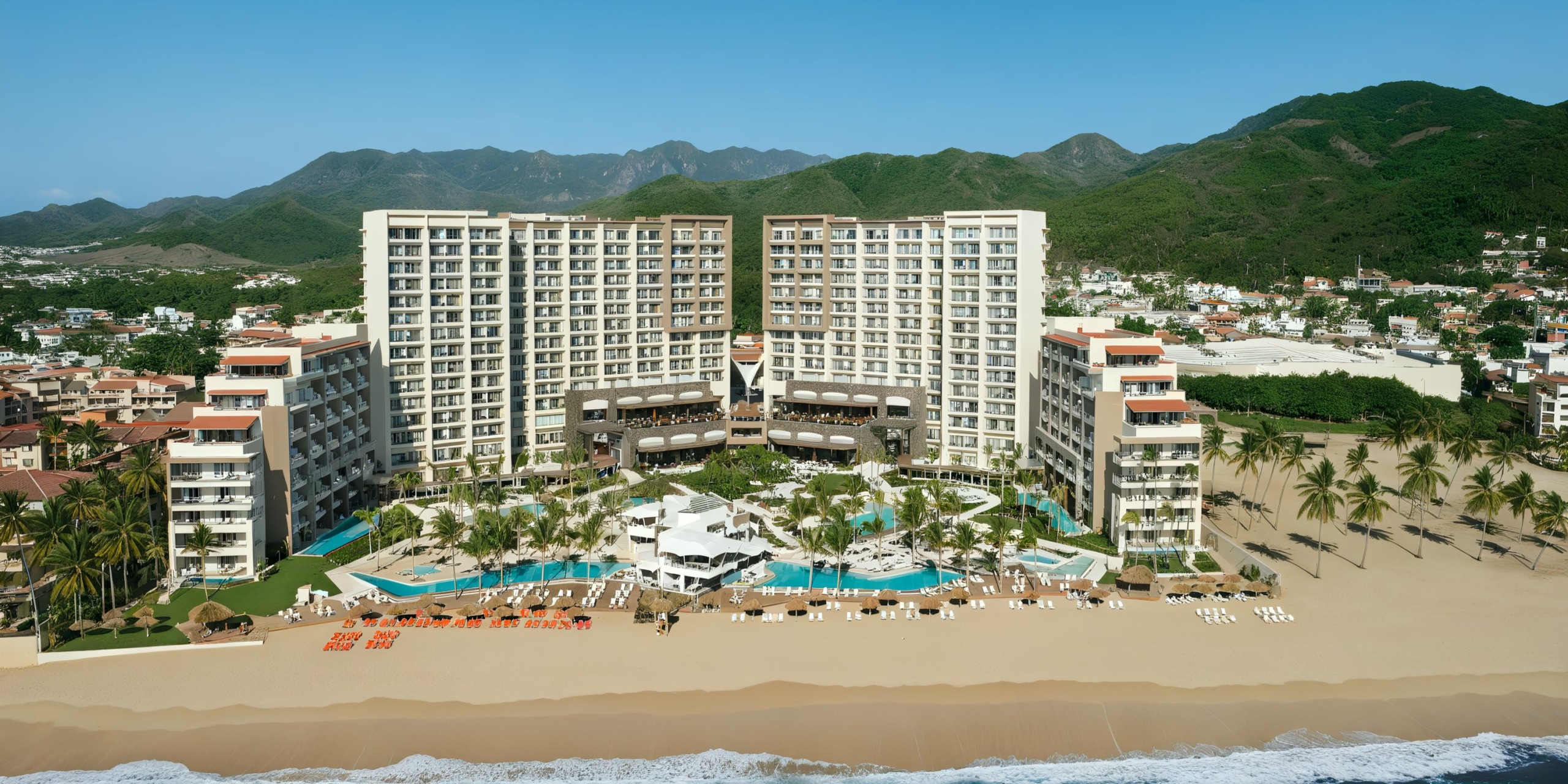 Secrets Vallarta Bay is a luxurious beachfront resort featuring two tall connected buildings nestled among lush palm trees, sparkling pools, and inviting lounge chairs. Stunning mountain views provide a breathtaking backdrop.