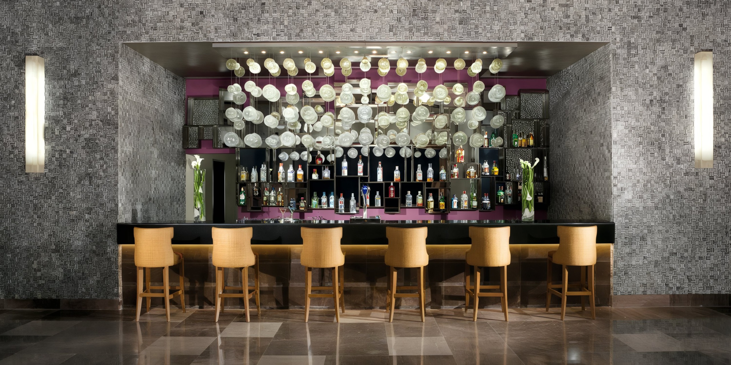 The Secrets Vallarta Bay features a contemporary bar complete with seven beige chairs, a purple backdrop, and shelves filled with bottles. Above the bar, there's a decorative wall adorned with white circular elements.