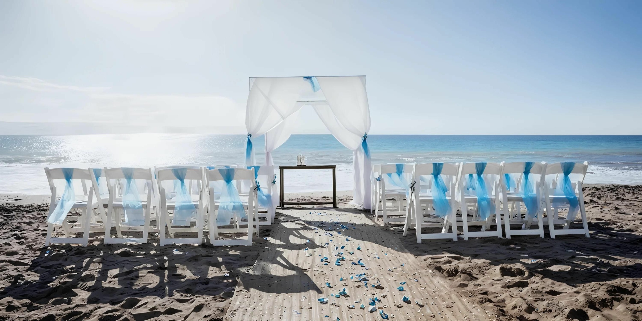 A beach wedding setup with white chairs and blue ribbons, a small table, and a white canopy facing the ocean. Blue rose petals are scattered along the sandy aisle.