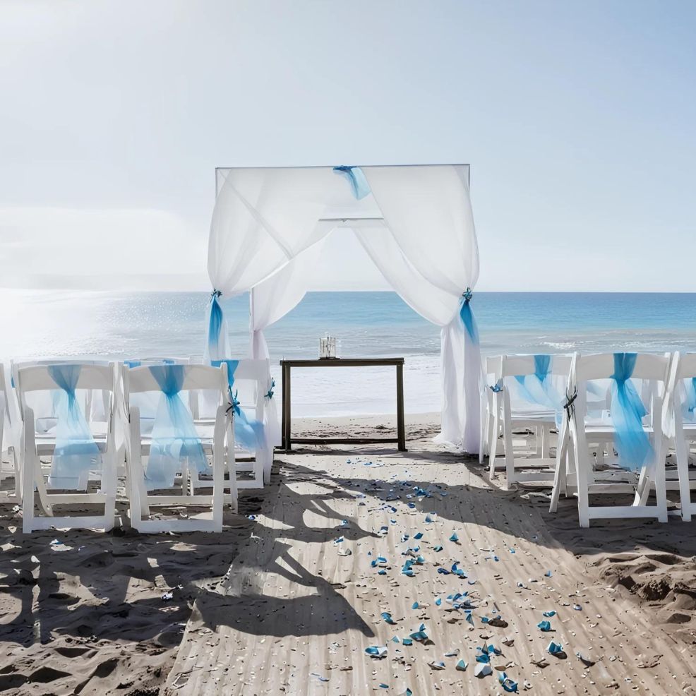 Beach wedding setup with white chairs adorned with blue sashes, a wooden walkway covered with blue and white flower petals, and a canopy overlooking the ocean.