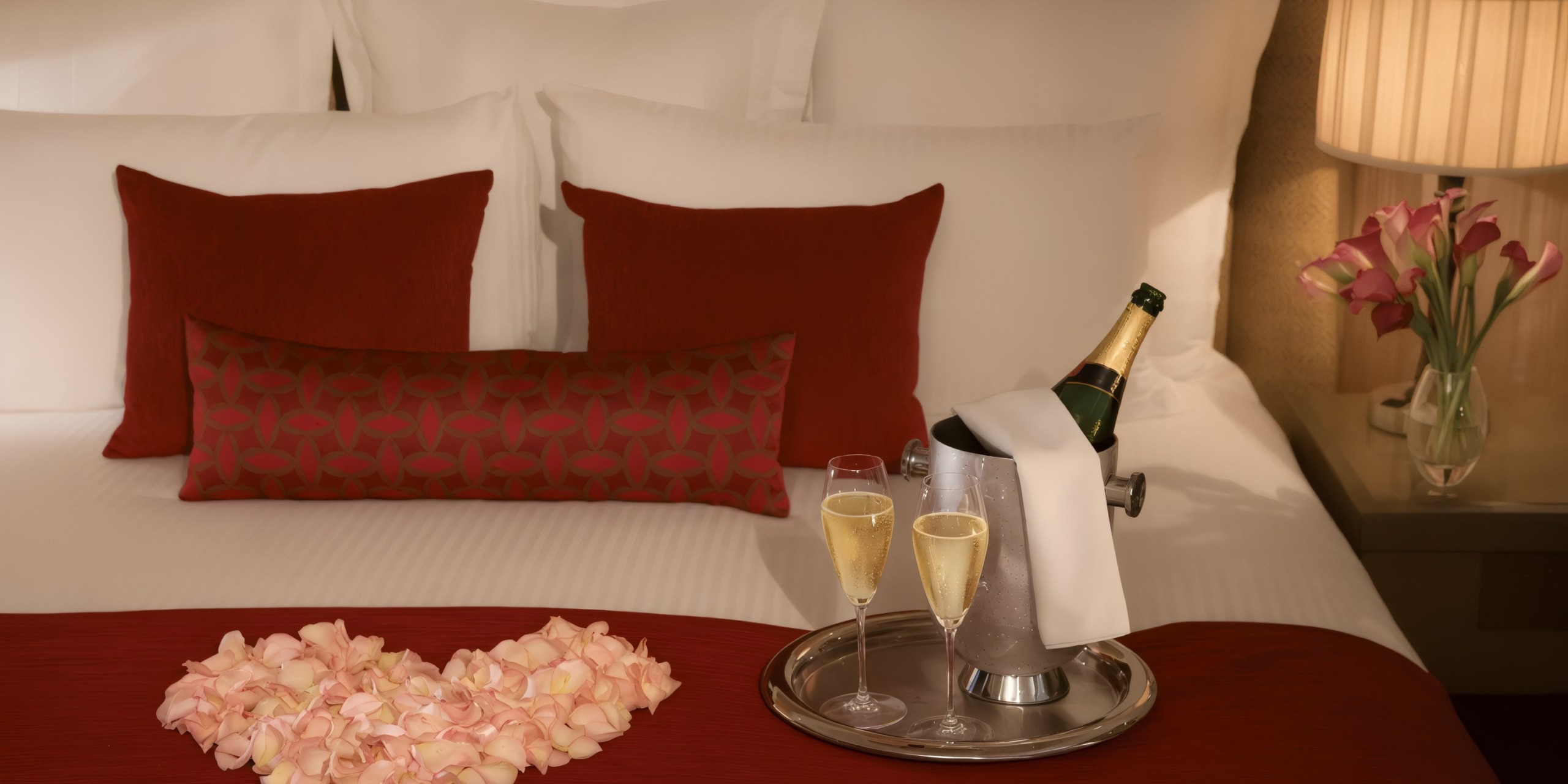The Secrets Vallarta Bay features a bed adorned with red and white pillows, highlighting a heart crafted from rose petals. Nearby is a tray holding an ice bucket with a champagne bottle and two glasses.