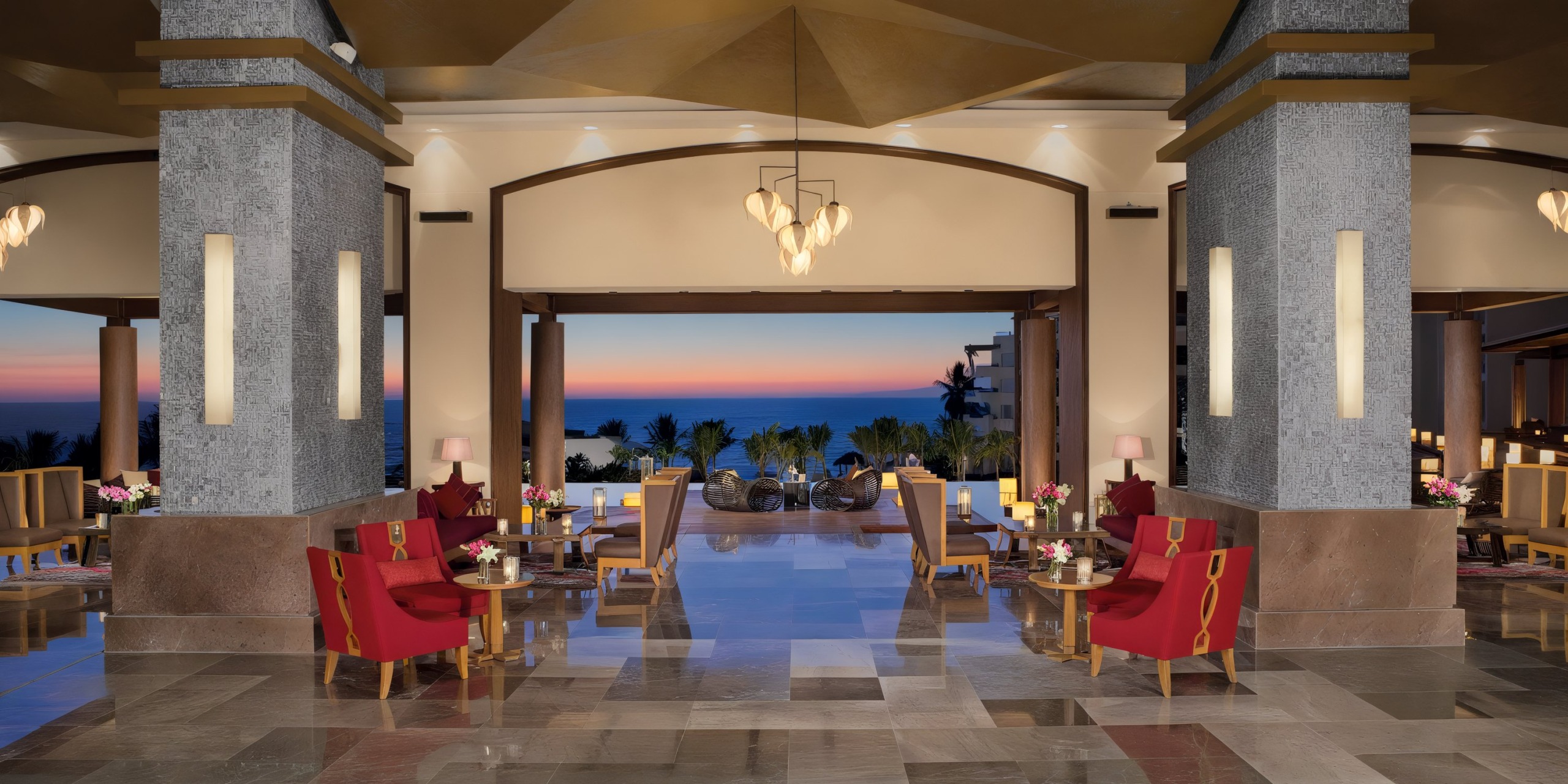 Experience the luxurious Secrets Vallarta Bay lobby, featuring elegant red seating, polished floors, and decorative lighting. Large windows reveal a stunning sunset view in the background.