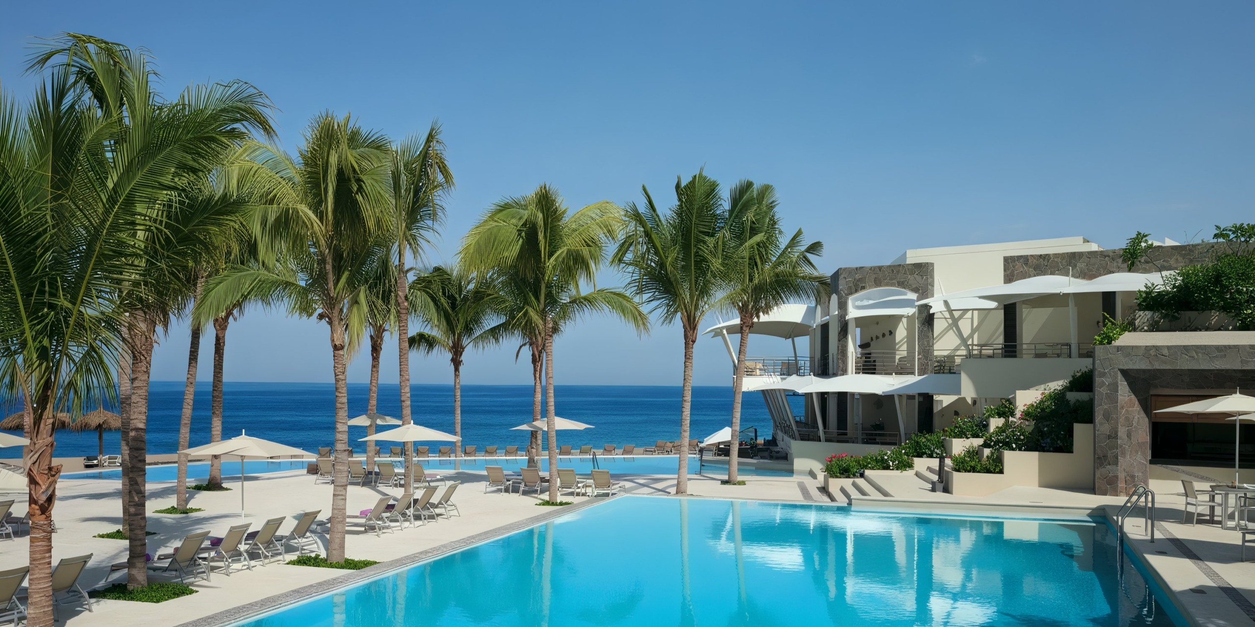 The Secrets Vallarta Bay boasts a pristine pool area adorned with palm trees, sun loungers, and umbrellas that overlooks a stunning bright blue ocean beneath a clear sky.
