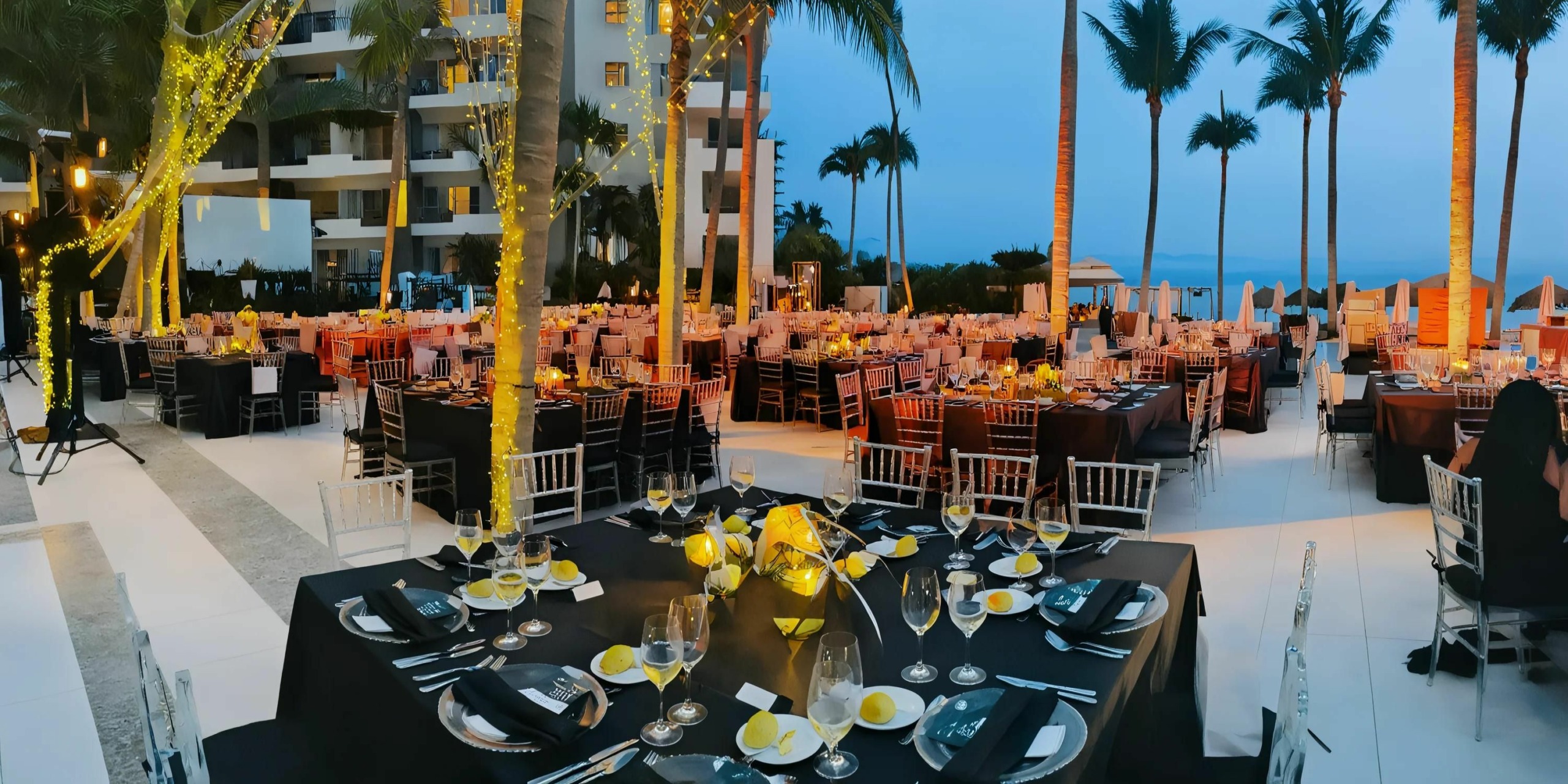 An outdoor evening banquet setup with tables and chairs arranged under palm trees adorned with string lights.