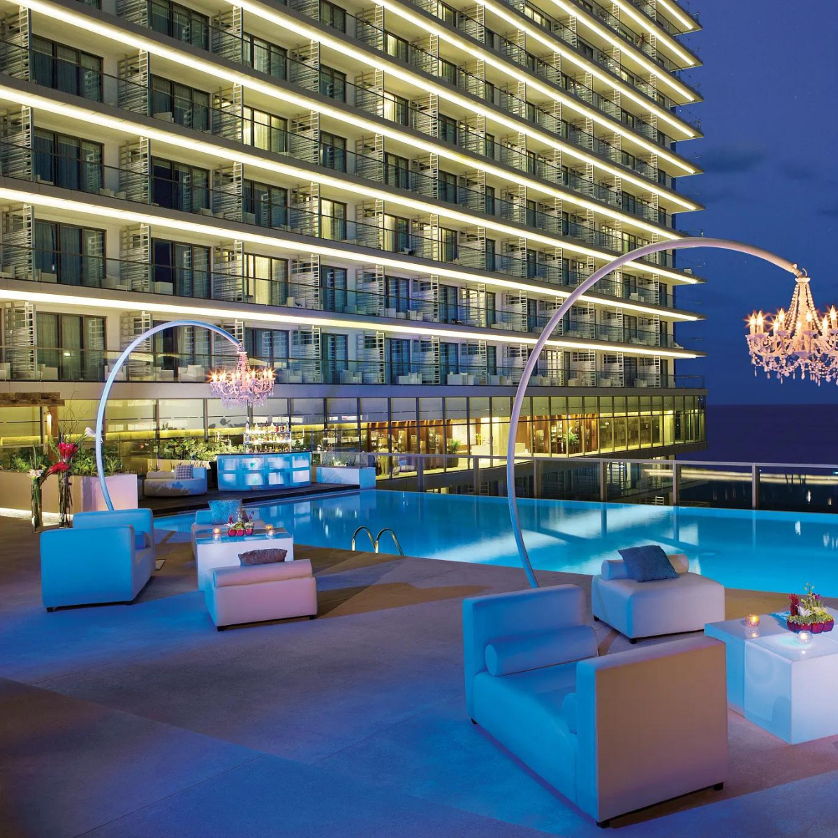 Secrets the Vine Cancun hotel poolside area at night with illuminated arches, a chandelier, modern seating, and a reflective water surface, perfect for destination weddings, with a multi-story building facade in the background