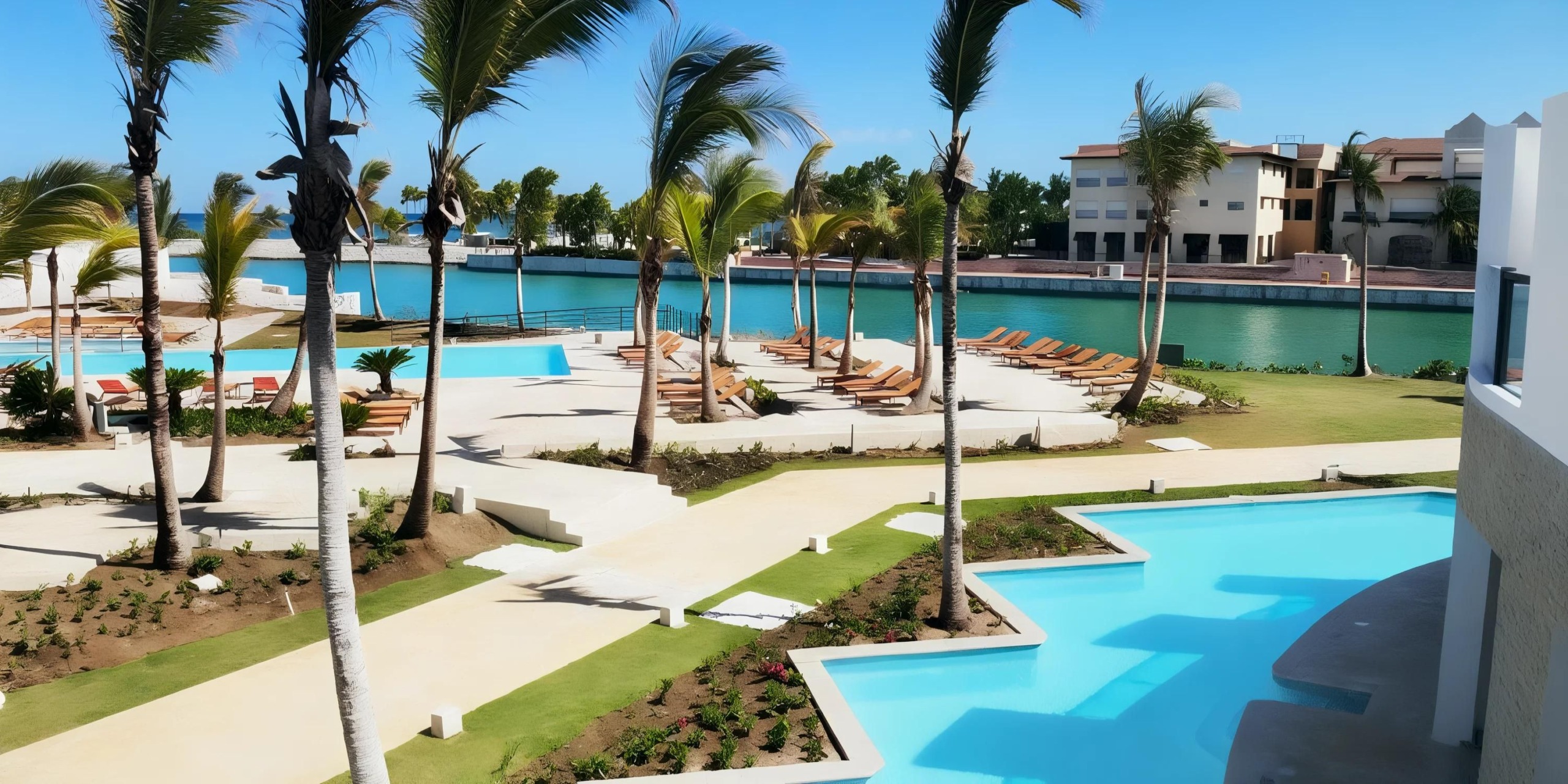 A resort scene featuring palm trees, multiple pools, sun loungers, and buildings along a tranquil waterfront under a clear blue sky.