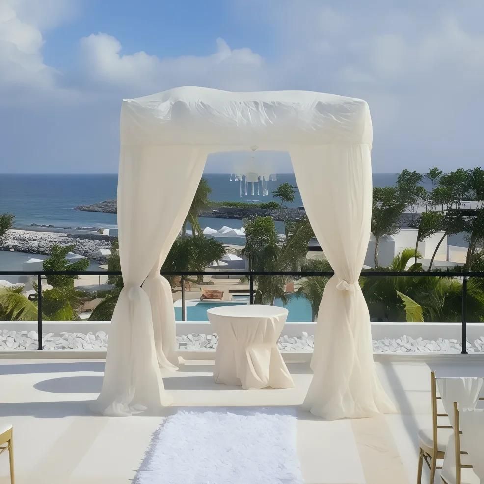 A white wedding arch with draped fabric stands on a terrace overlooking the ocean. There's a small round table beneath the arch and a white aisle runner leading to it.