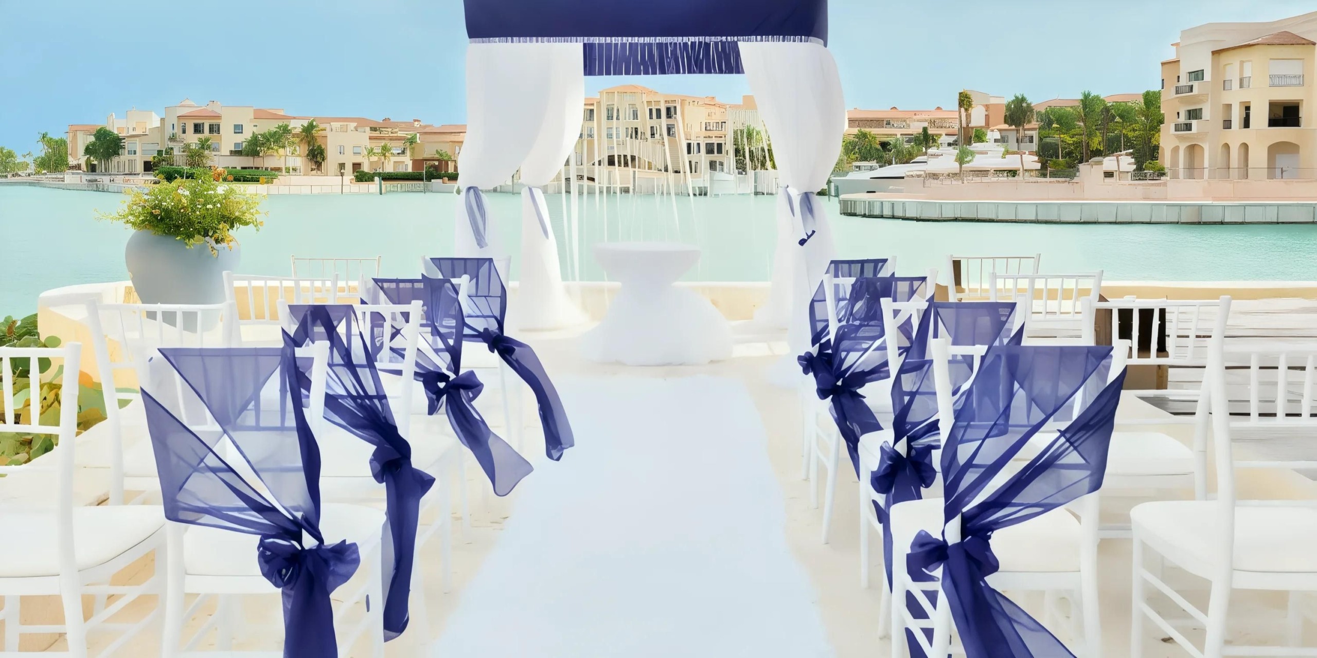Outdoor wedding setup with white chairs adorned with blue sashes, a white aisle runner, and an archway overlooking water and buildings in the background.