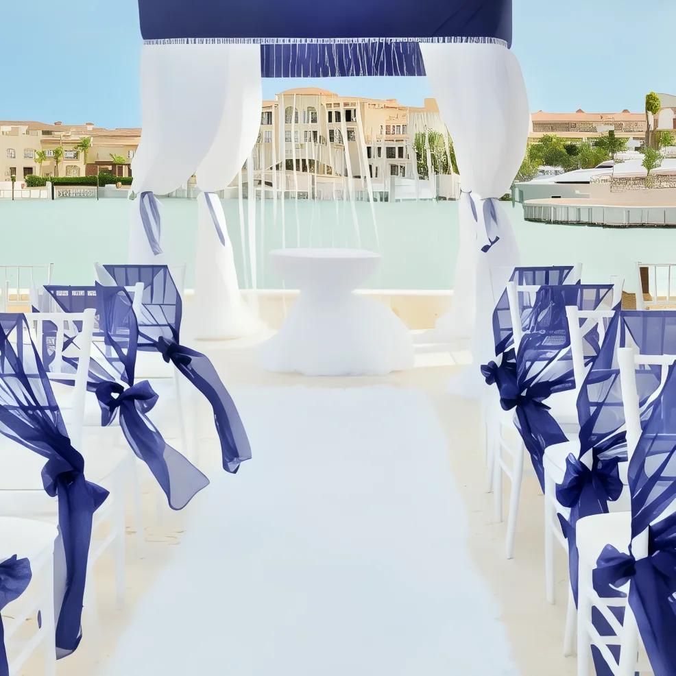 Outdoor wedding ceremony setup with white chairs adorned with navy blue bows. A white aisle leads to an altar overlooking water and buildings.