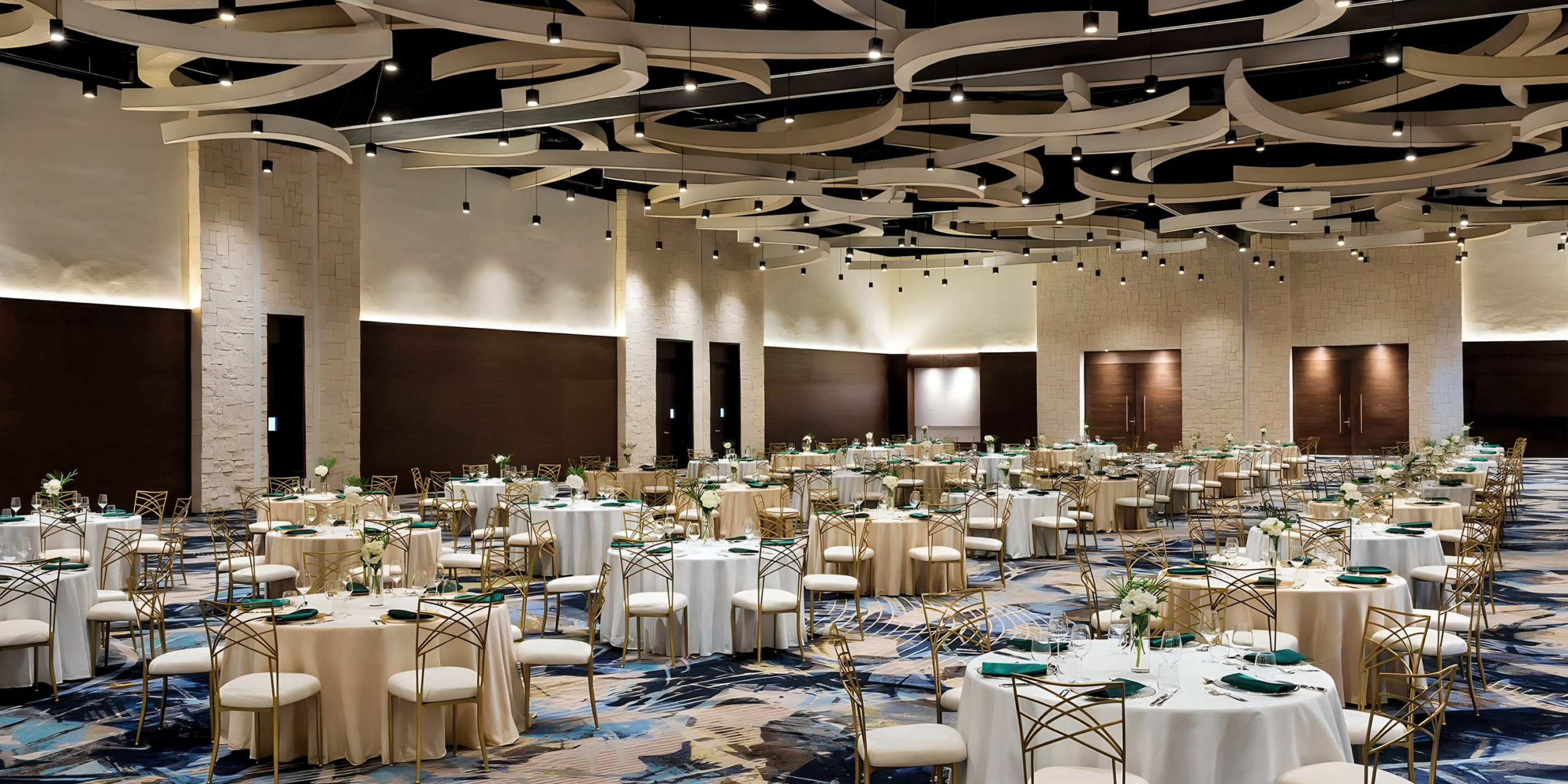 Elegant banquet hall set up for destination weddings with round tables, each covered with cream tablecloths and decorated with green centerpieces, under a contemporary white ceiling design.