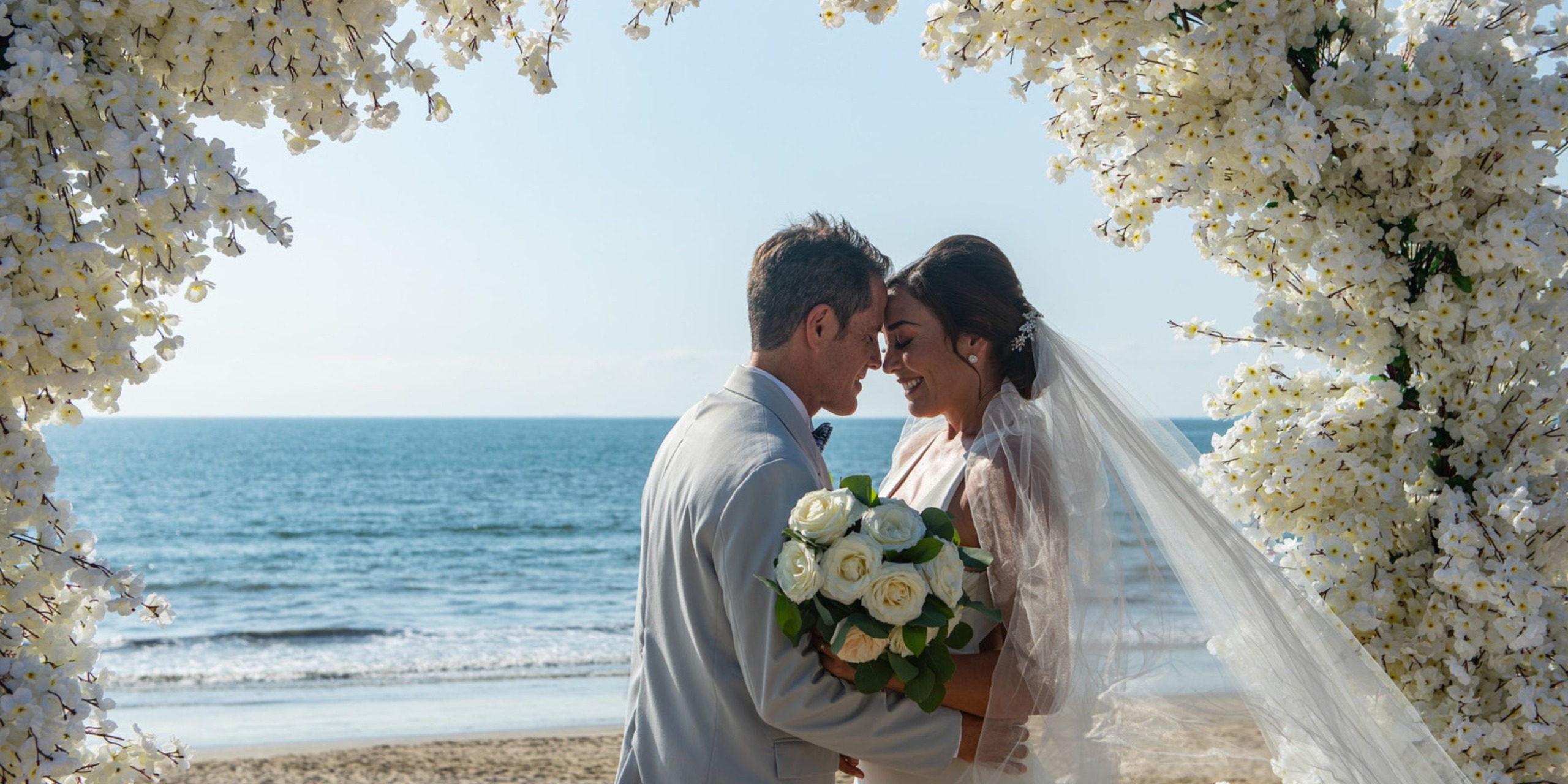 The bride and groom are beaming under a floral arch on the beach, celebrating their beautiful destination wedding.