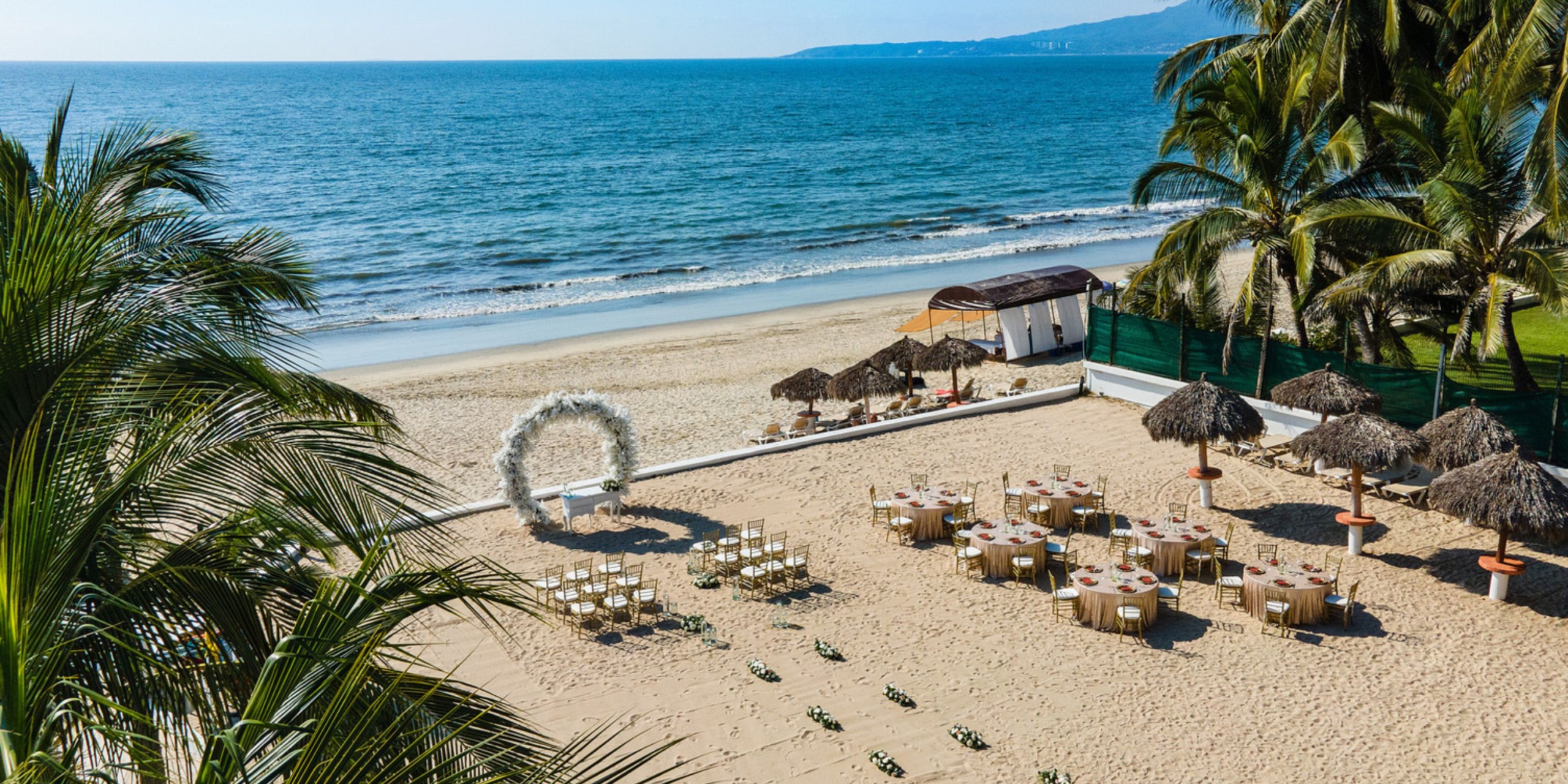 A destination wedding setup on the beach featuring a charming arch, elegantly arranged chairs, and tables shaded by umbrellas on the sandy shore.
