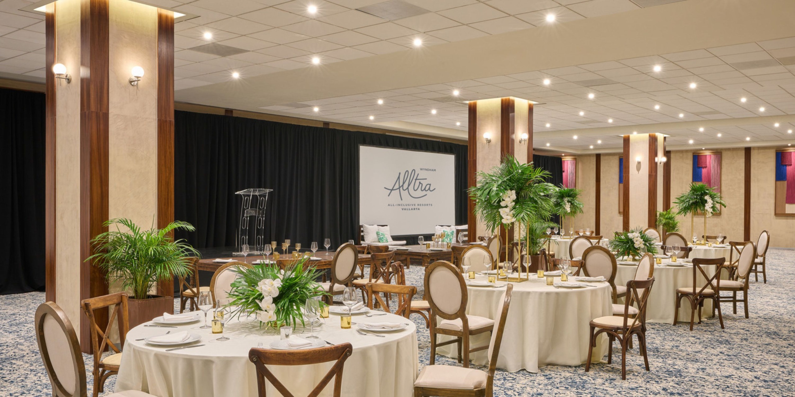 Wyndham Alltra Vallarta offers a chic banquet hall, perfect for a destination wedding, featuring round tables adorned with elegant floral decor and a stage complete with a podium.