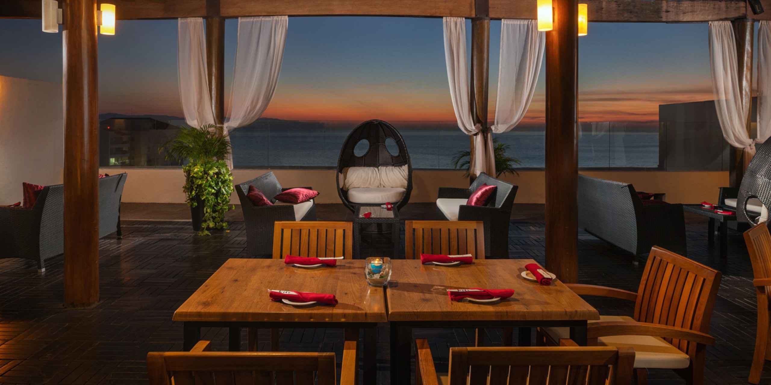 A rooftop dining area featuring a wooden table and chairs offers a breathtaking ocean view at sunset, perfect for a destination wedding.