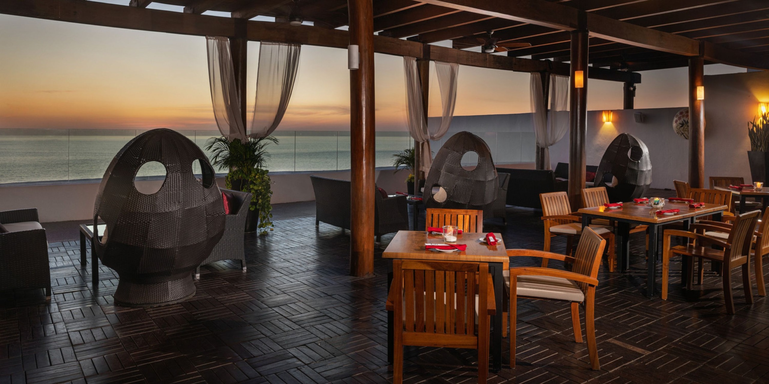 A romantic outdoor dining area featuring wooden tables and chairs provides a picturesque setting for a destination wedding, all while overlooking the ocean at sunset.
