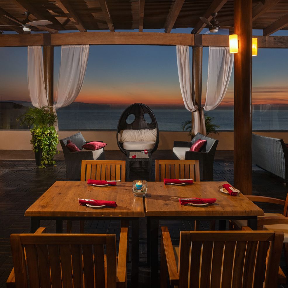 A covered outdoor patio features a dining table and seating, offering a breathtaking ocean view at sunset—perfect for an unforgettable destination wedding setting.