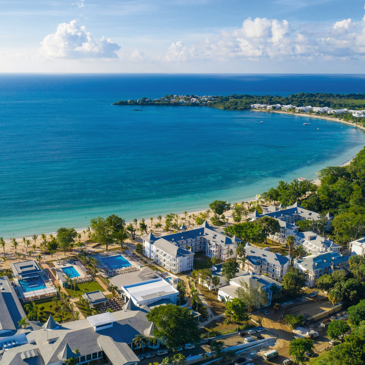 Aerial view of Hotel Riu Palace Tropical Bay, a popular destination for weddings, with white buildings, swimming pools, and palm trees along a sandy beach adjacent to a clear blue sea.