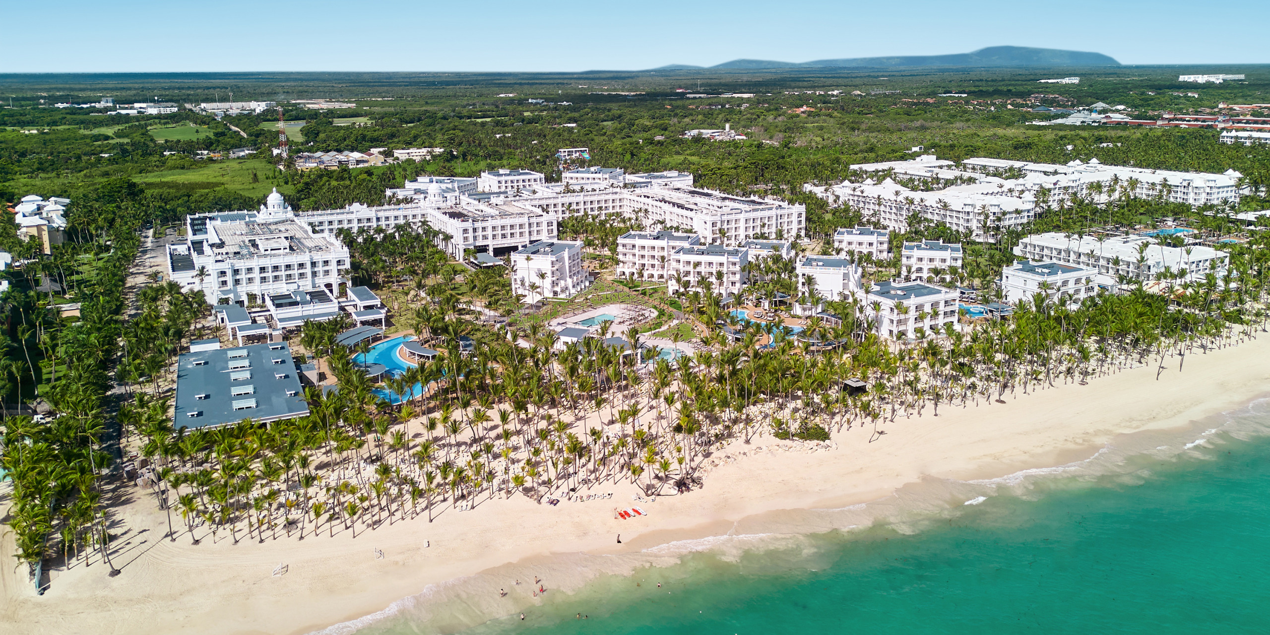 Aerial view of Hotel Riu Palace Bavaro, a spacious beachfront resort featuring multiple buildings, pools, and palm trees, situated next to the turquoise ocean waters and a sandy beach.