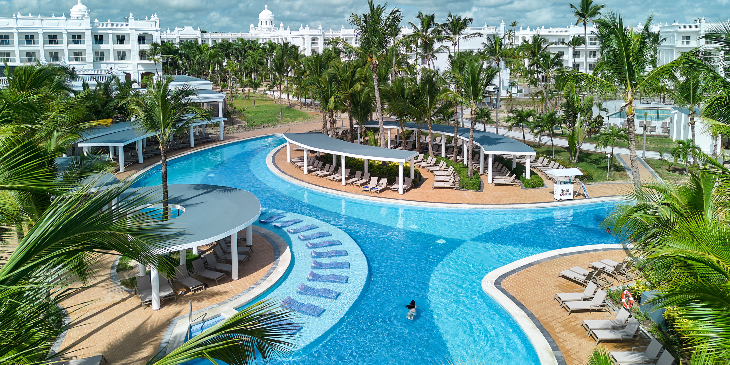 Aerial view of the Hotel Riu Palace Bavaro's luxury resort pool featuring curved sun loungers, palm trees, and white buildings in the background.