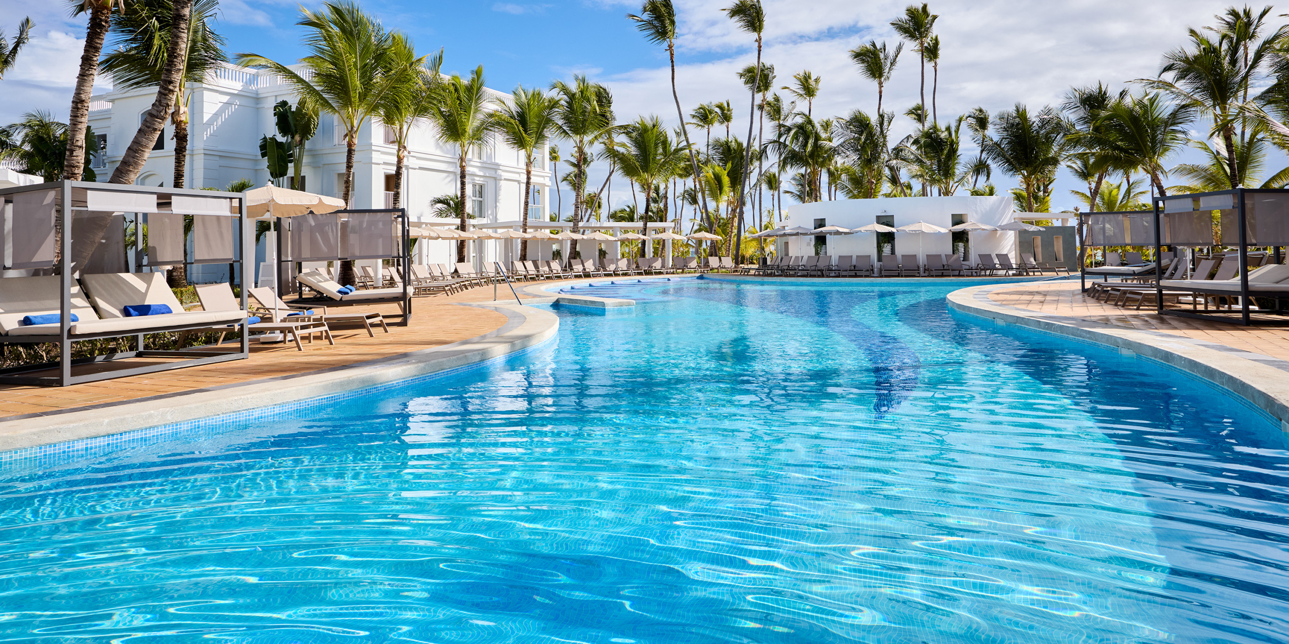 Hotel Riu Palace Bavaro features an outdoor swimming pool encircled by lounge chairs and palm trees, set against a white building backdrop under a partly cloudy sky.