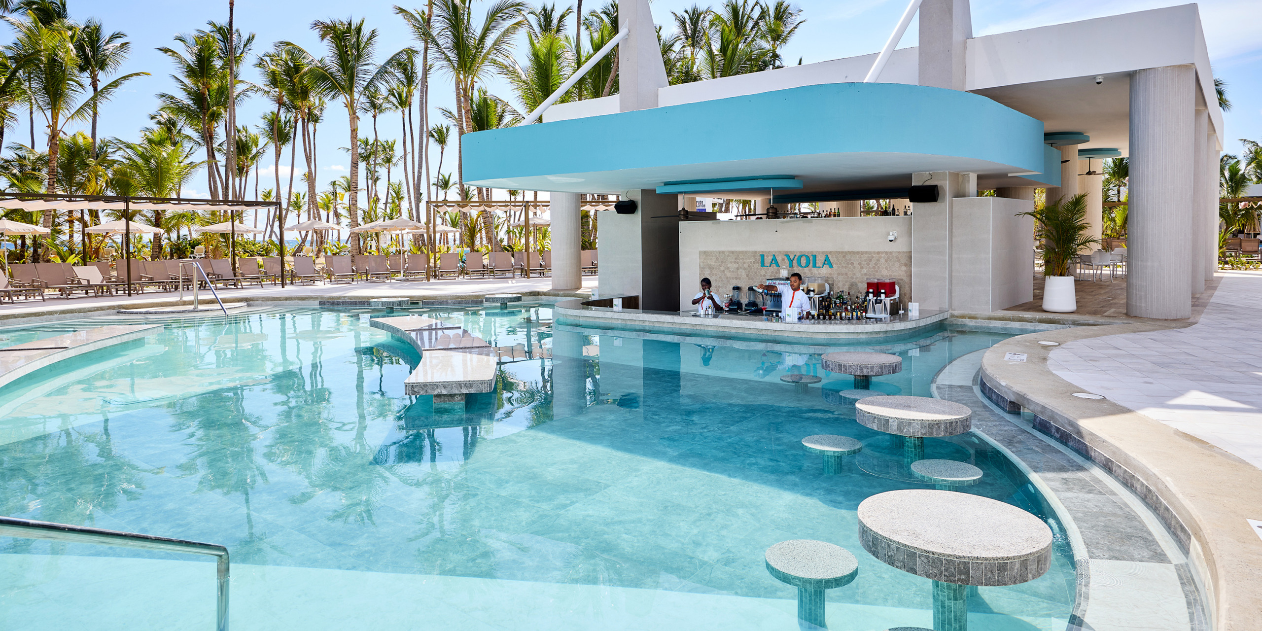 The Hotel Riu Palace Bavaro features an outdoor pool with a swim-up bar named "La Yola," complete with bar stools in the water and surrounded by palm trees and lounge chairs.