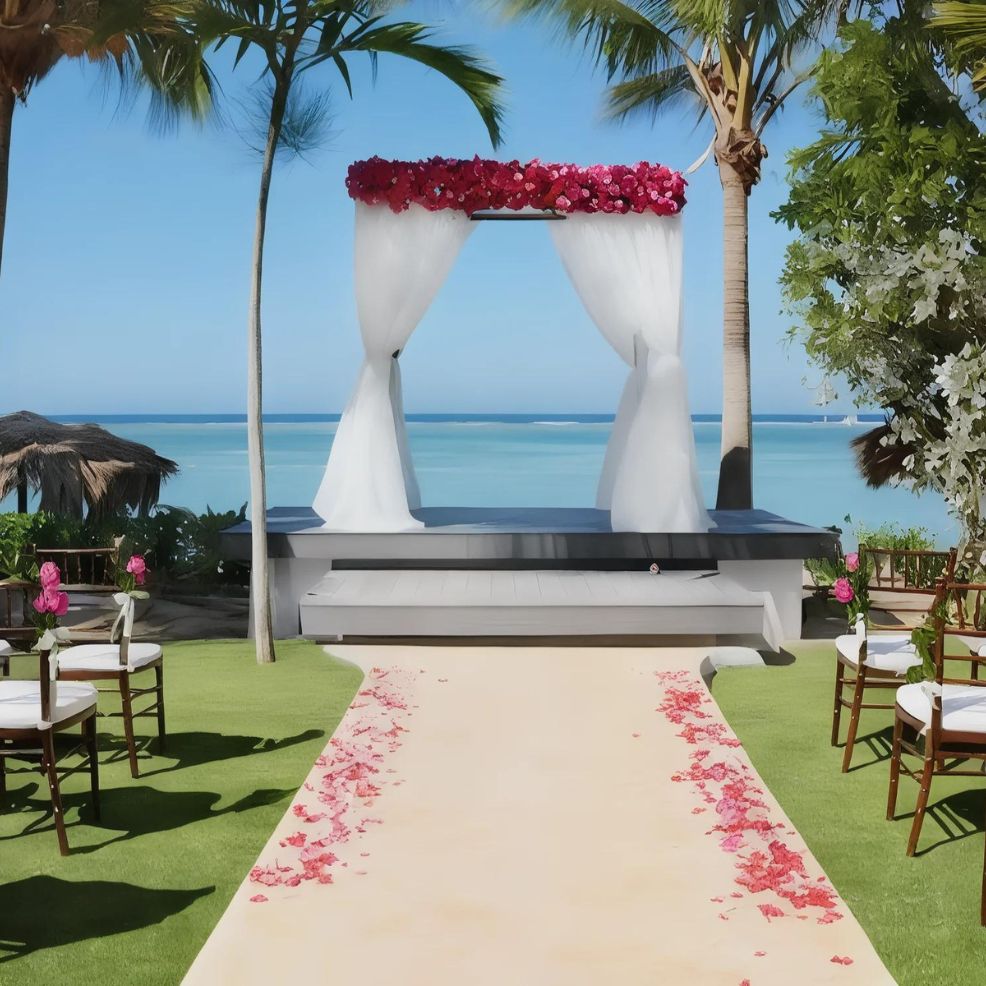 An outdoor wedding setup with an aisle lined with pink petals, leading to an altar decorated with white drapes and pink flowers. The background features serene ocean views and palm trees.
