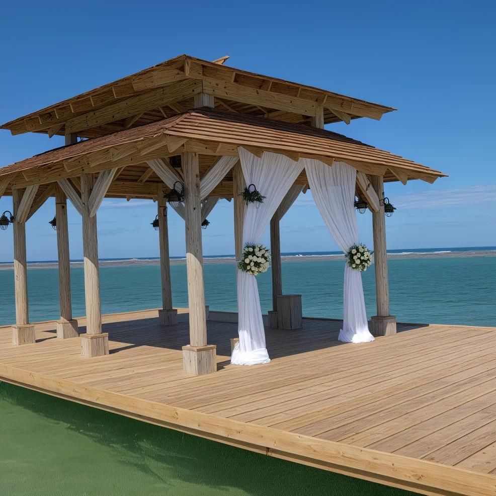 A wooden gazebo with white curtains and floral decorations stands on a wooden deck over the water with a clear blue sky in the background.