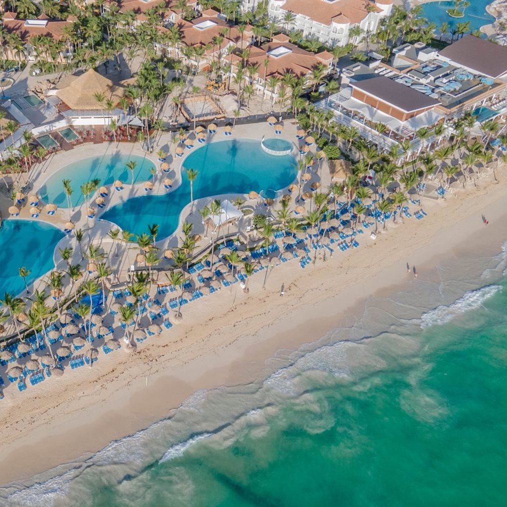 A bird's-eye view of Grand Punta Cana reveals a stunning wedding destination, complete with pools, palm trees, and sun loungers lining the picturesque shore.