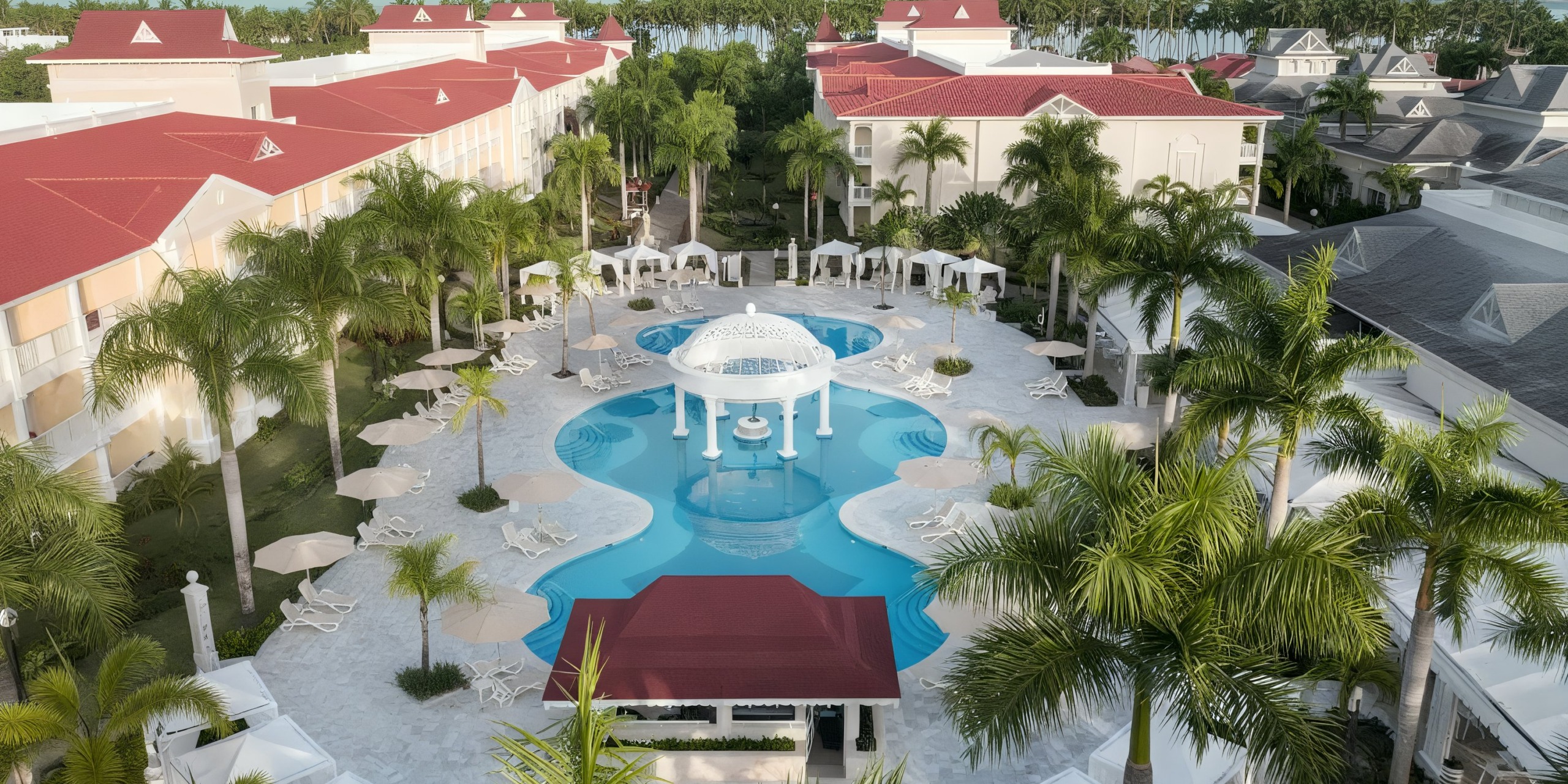 Aerial view of the Bahia Principe Luxury Bouganville pool area, featuring palm trees, white loungers, and a circular pavilion. The hotel buildings are adorned with red roofs and beige walls.
