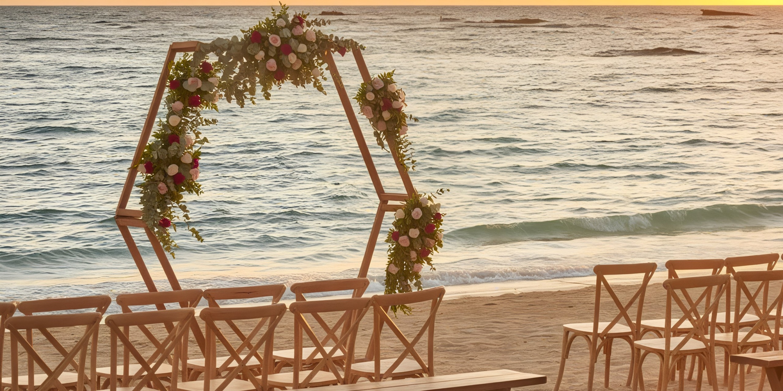 A hexagonal floral arch and rows of chairs are arranged on the beach at Bahia Principe Luxury Bouganville, with ocean waves and a sunset creating a stunning backdrop.