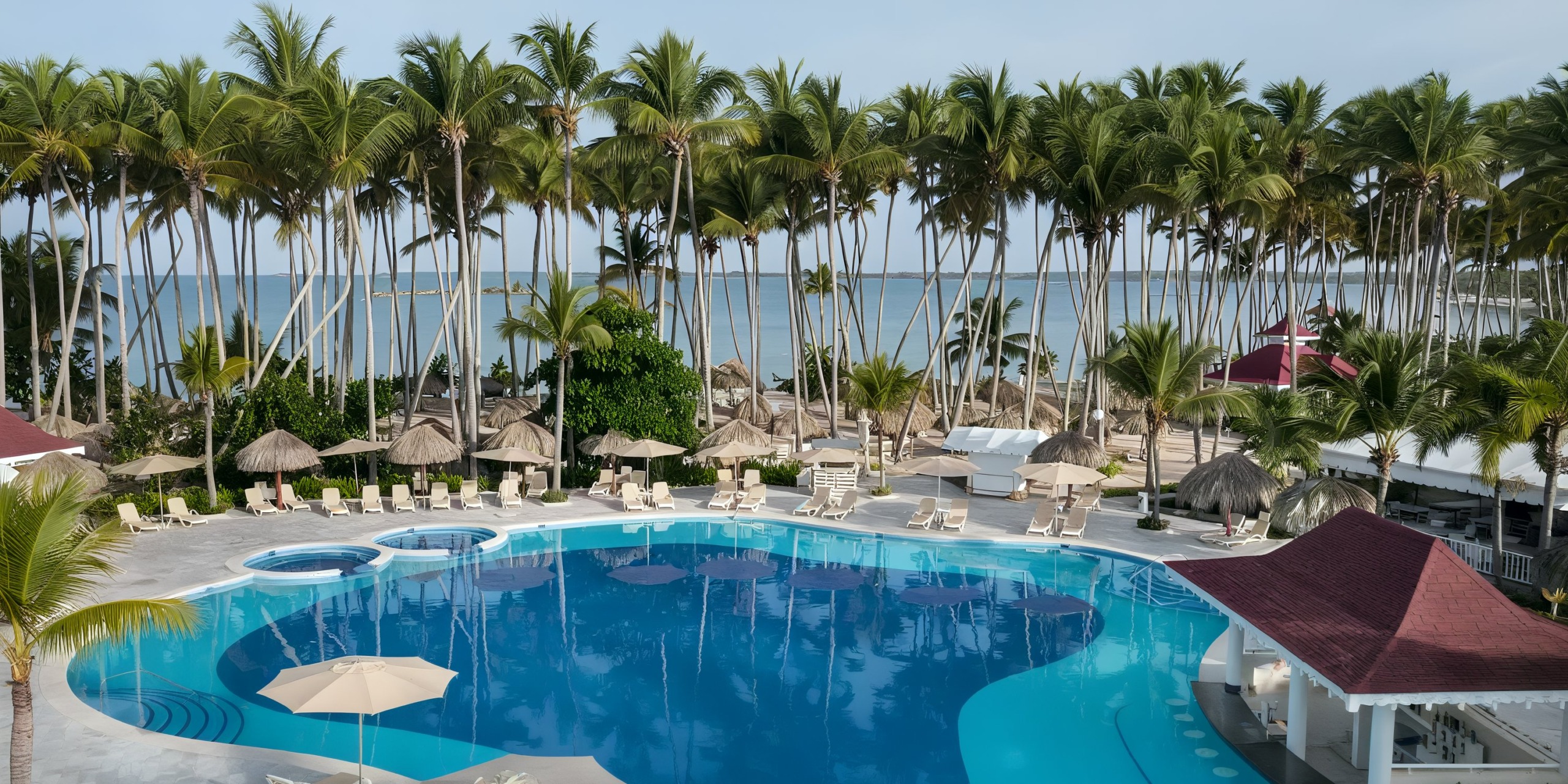 A picturesque view at Bahia Principe Luxury Bouganville features a spacious pool encircled by palm trees and lounge chairs, with the ocean visible in the backdrop.