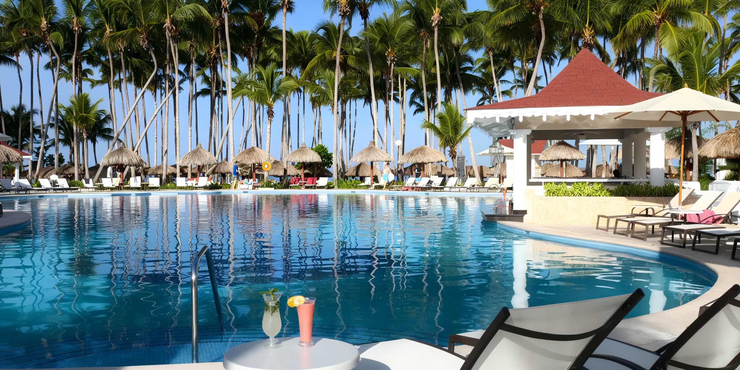 Experience the serene pool area at Bahia Principe Luxury Bouganville, where lounge chairs surround a thatched-roof bar and palm trees sway under a clear blue sky. Refreshing drinks are set on a nearby table, completing the idyllic scene.