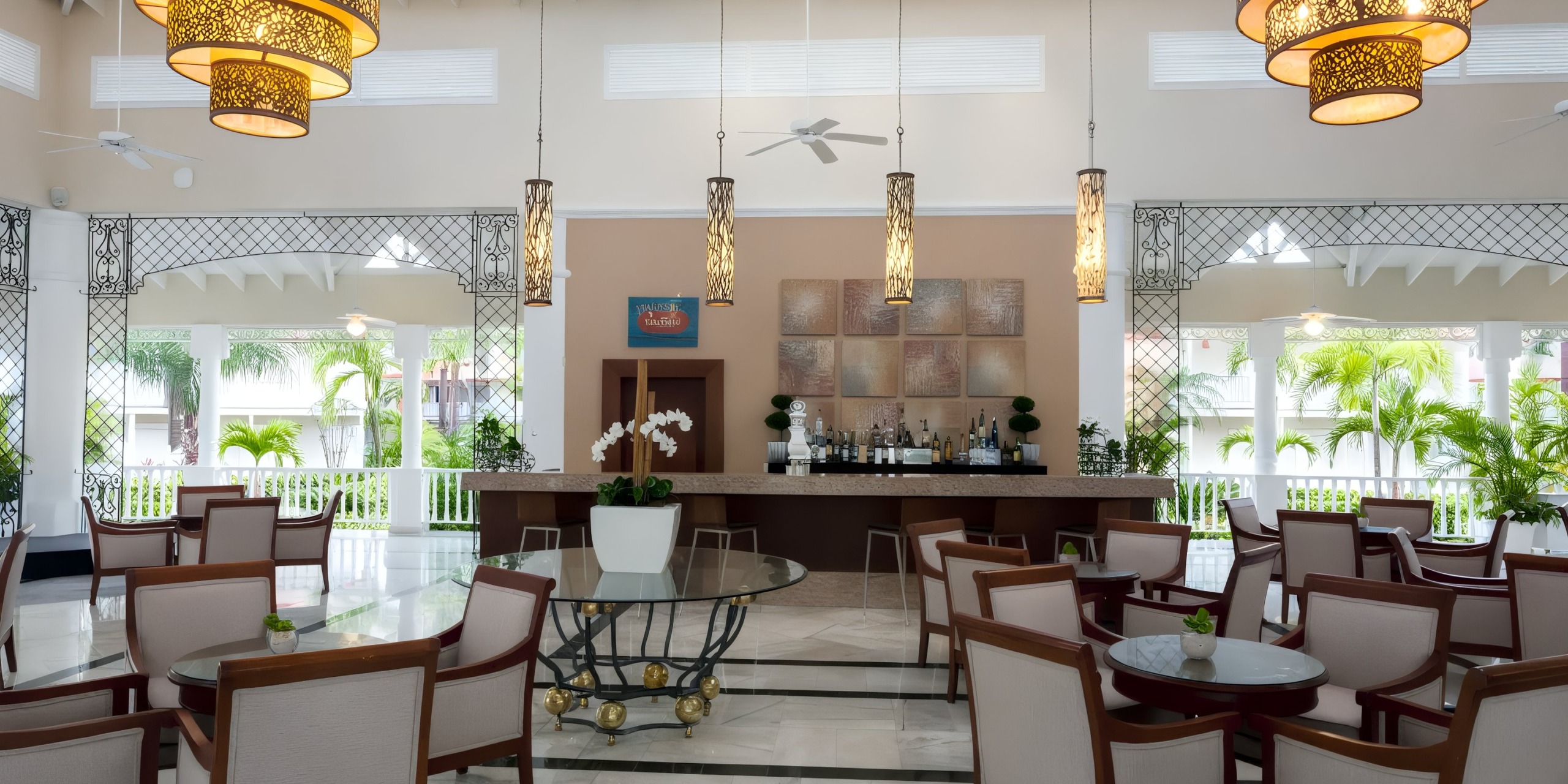 The Bahia Principe Luxury Bouganville offers a spacious hotel bar with modern decor, showcasing a round glass table adorned with a potted plant, hanging lights, wooden chairs, and open views of palm trees through the windows.