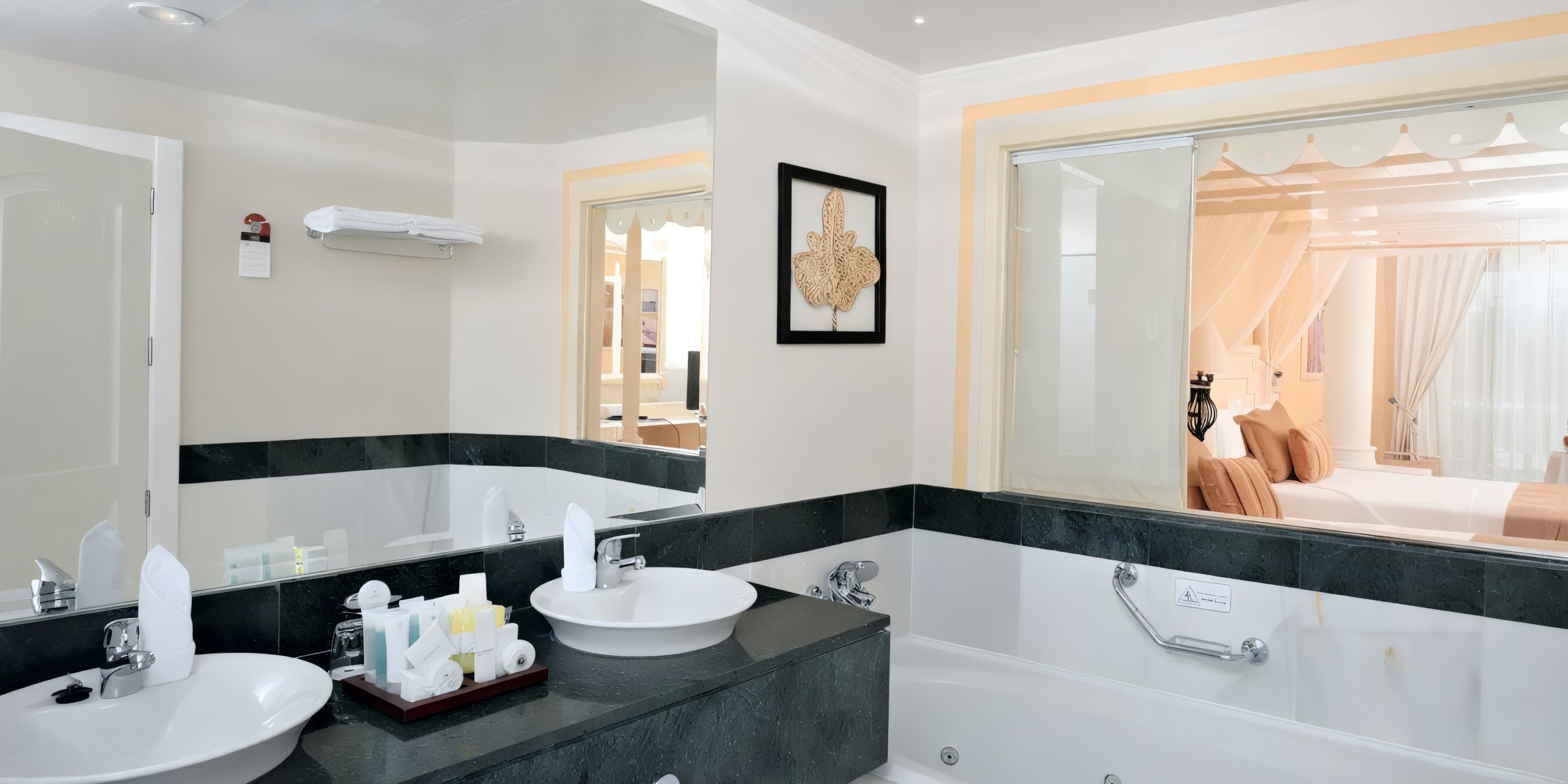 The luxurious bathroom at Bahia Principe Luxury Bouganville includes dual sinks, a bathtub, a wall-mounted mirror, and opens to the bedroom adorned with sheer curtains and plush pillows.