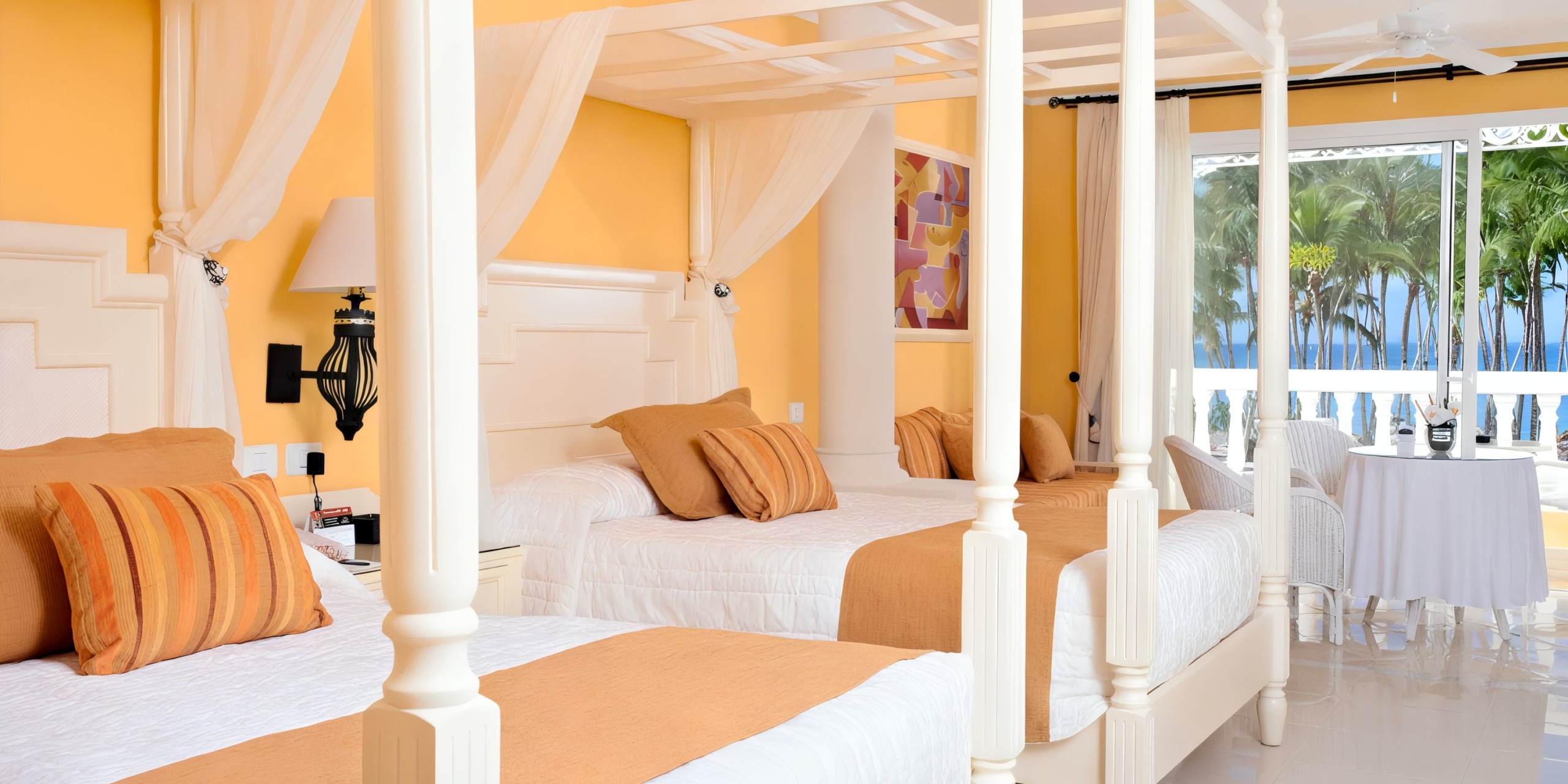 Experience a vibrant room at the Bahia Principe Luxury Bouganville, featuring two canopy beds, orange and white decor, a cozy table with chairs, and a private balcony offering stunning views of palm trees and the ocean.
