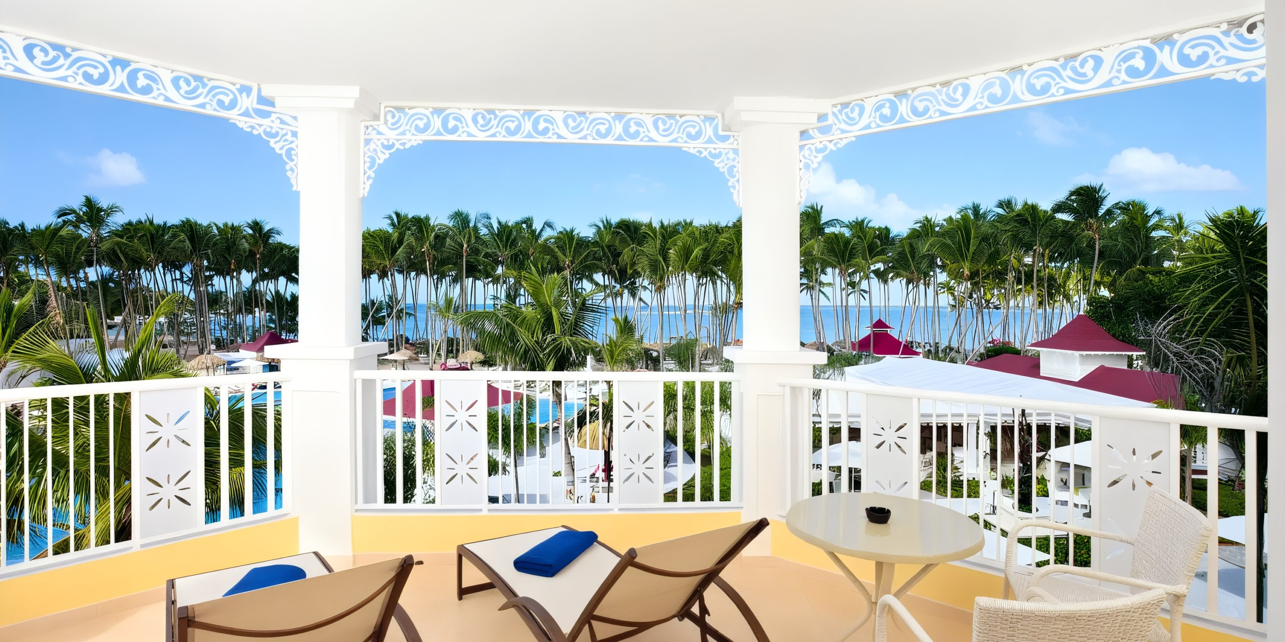 Enjoy a balcony at the Bahia Principe Luxury Bouganville, where you can relax in two lounge chairs along with a round table. The view features a resort landscape adorned with palm trees, a tranquil pool, and the ocean stretching into the background.