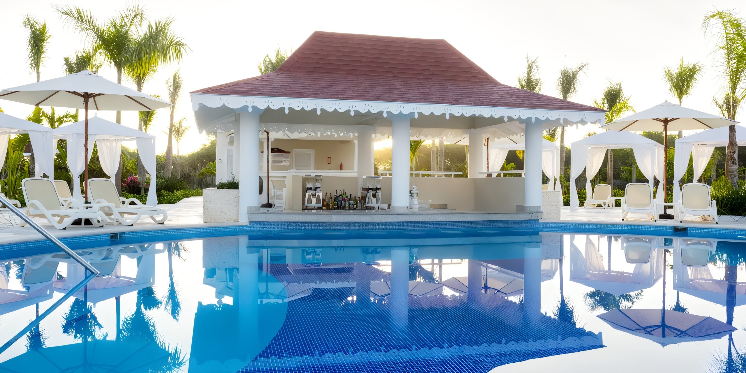 At the Bahia Principe Luxury Bouganville, a poolside bar featuring a red roof and white pillars is encircled by sun loungers and umbrellas, mirroring beautifully in the clear blue water beneath a bright sky.