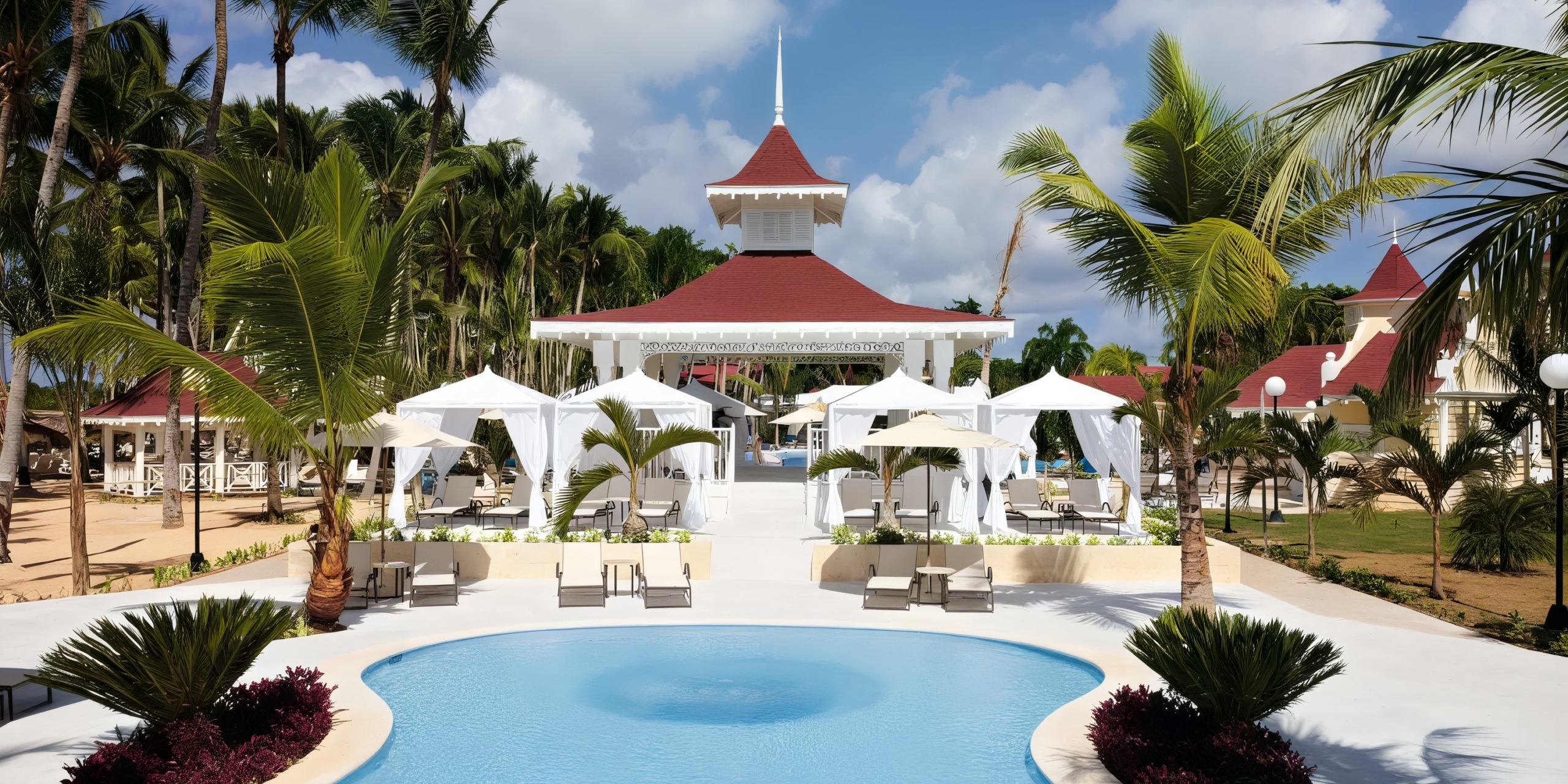 The pool area at Bahia Principe Luxury Bouganville features white cabanas, palm trees, and a building with a red roof beneath a partly cloudy sky.