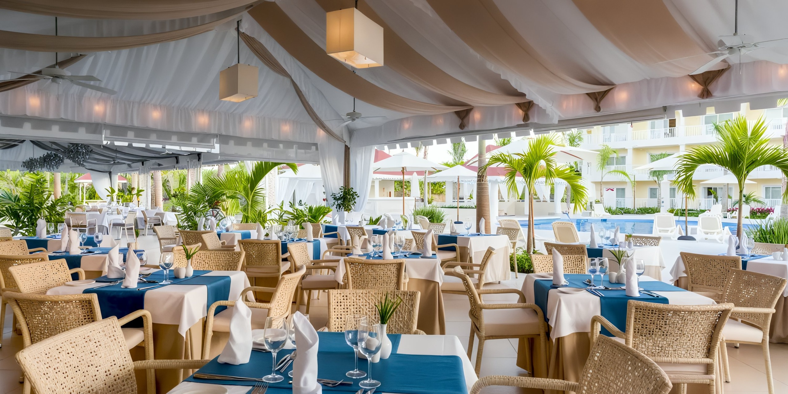 The open-air dining area at the Bahia Principe Luxury Bouganville is elegantly arranged with tables adorned in blue and white linens, complemented by glassware and napkins, all set beneath a charming canopy surrounded by lush plants, with a serene pool area in the background.
