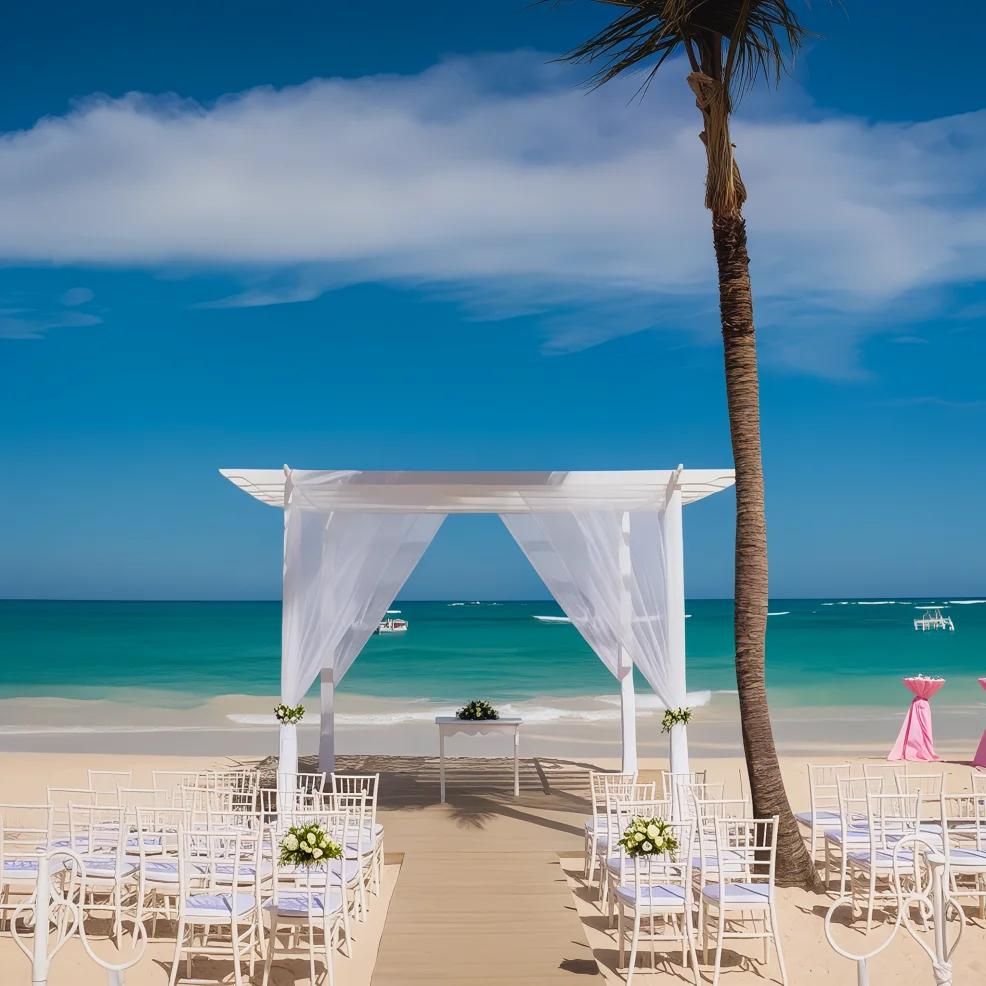 A beach wedding setup with a white canopy, chairs, and floral decorations. The ocean and a palm tree are in the background under a blue sky.