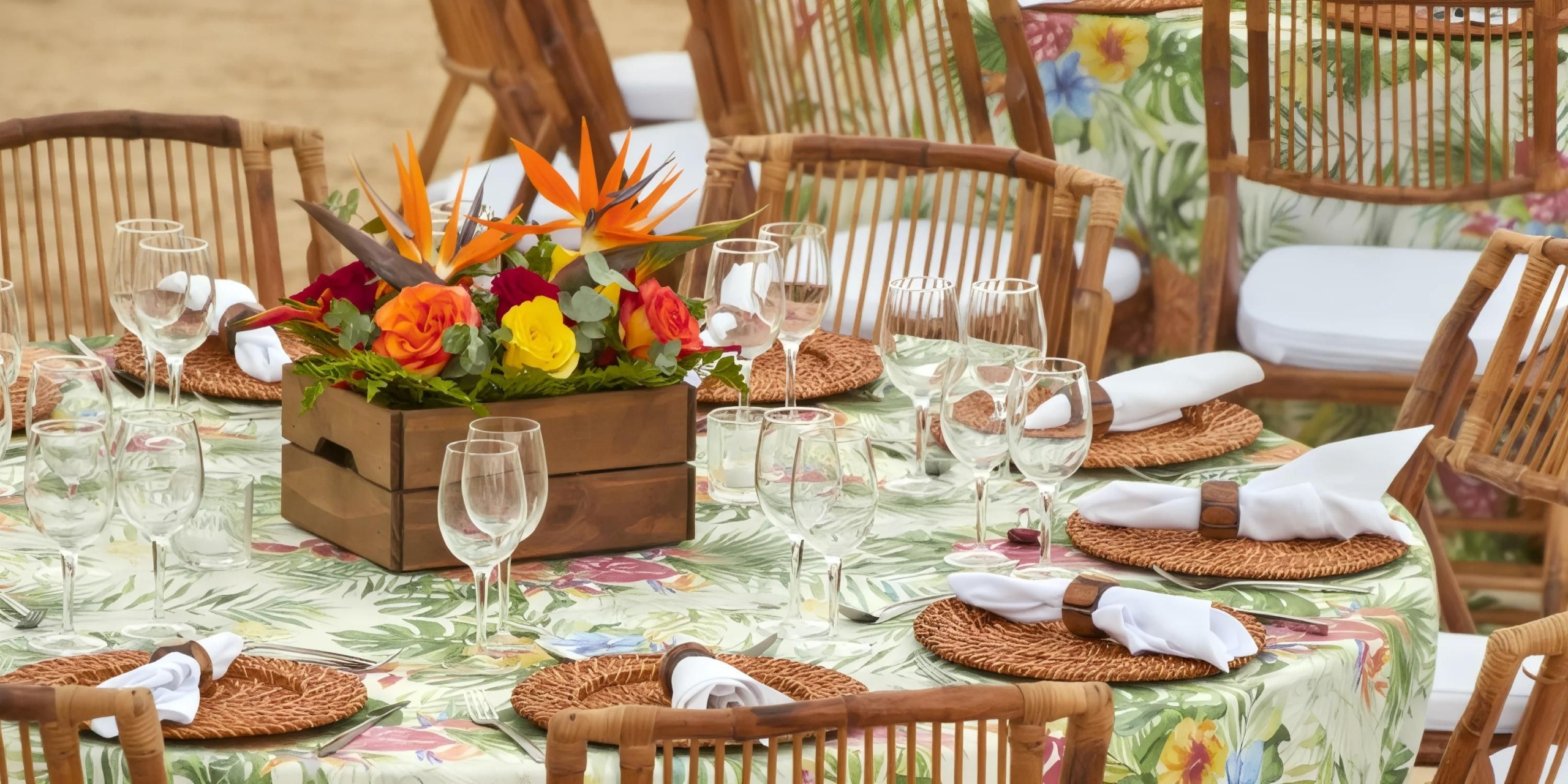 A round table with floral-patterned tablecloth, set with rattan placemats, white napkins, and clear glasses. A wooden box with colorful flowers serves as a centerpiece.