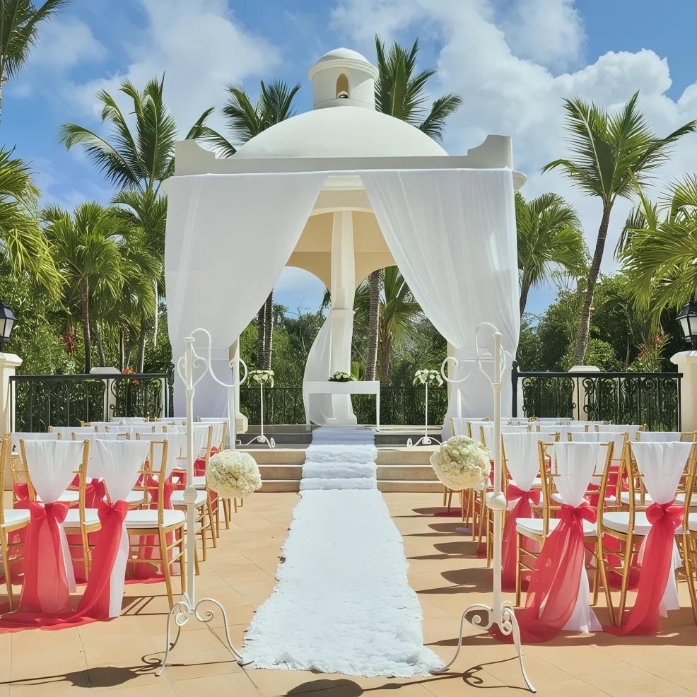 Outdoor wedding venue with a white gazebo, draped fabric, and a white aisle runner. Gold chairs with red sashes are arranged in rows under a blue sky with palm trees in the background.