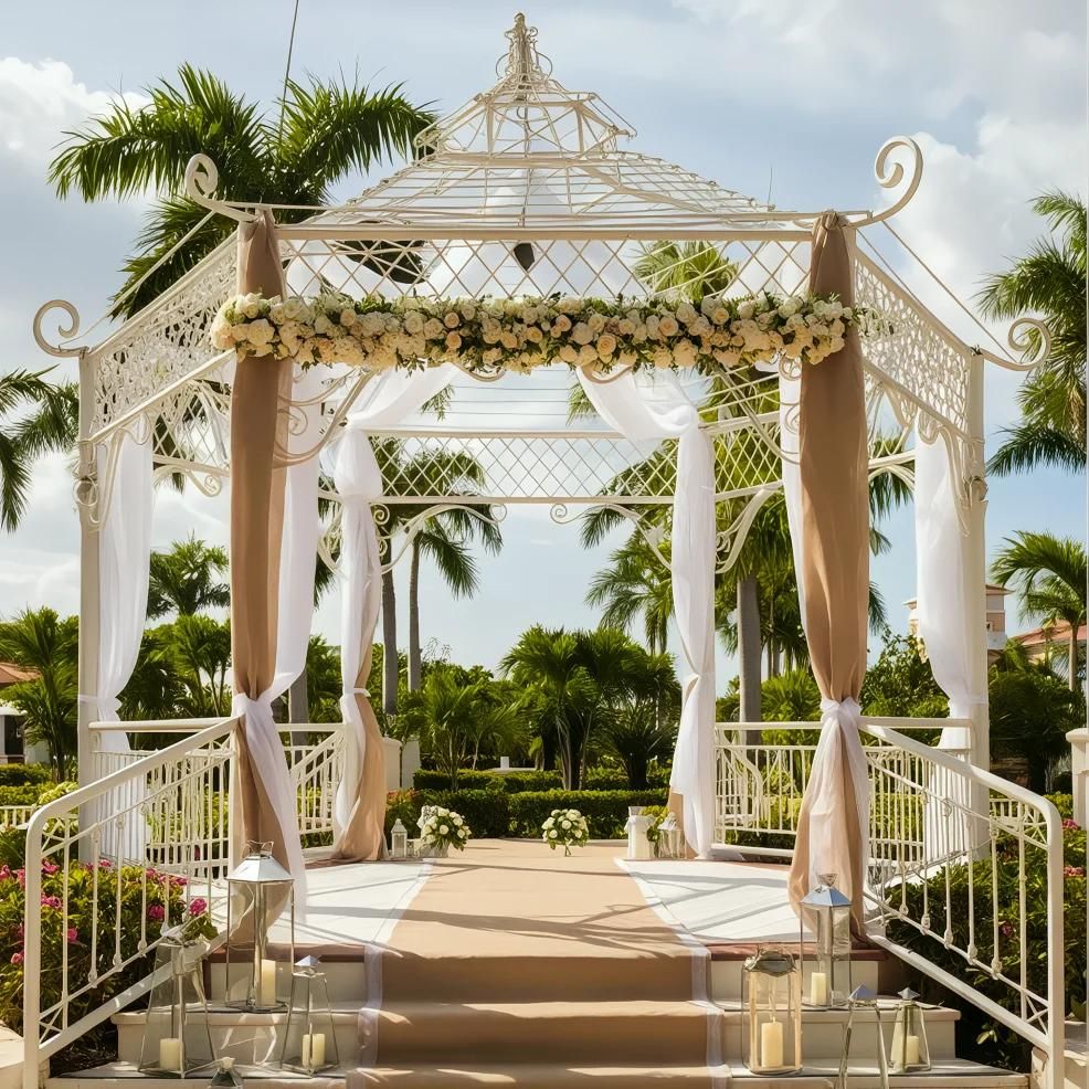 A white gazebo decorated with flowers and draped fabric stands on a sunny day, surrounded by palm trees and greenery.