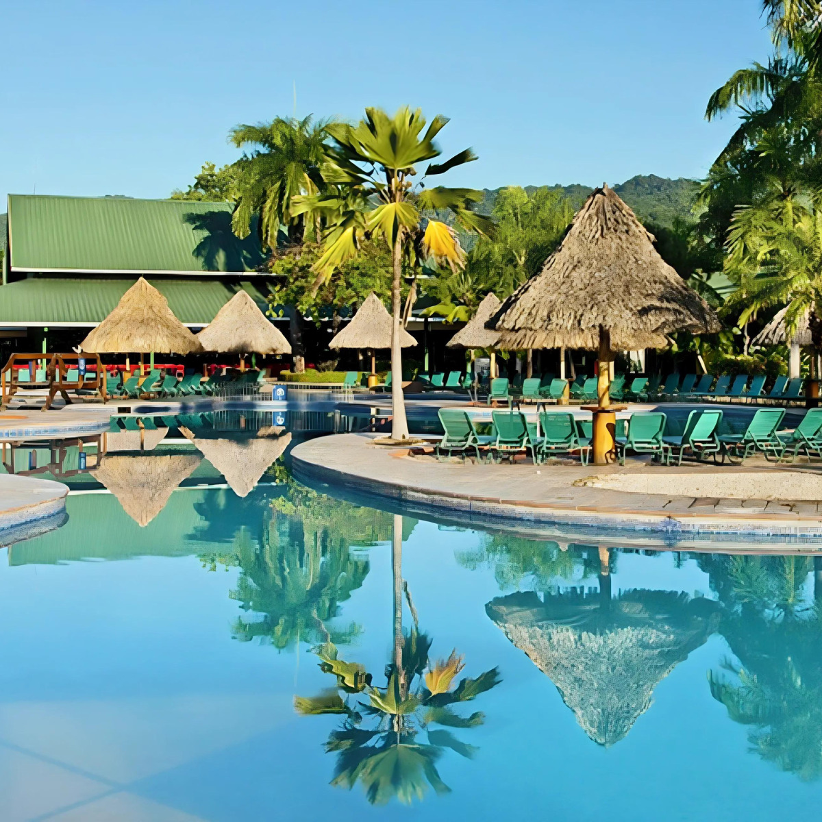 A Barcelo Tambor destination wedding resort pool with palm trees, thatched umbrellas, and loungers, reflecting in the calm water under a clear sky.