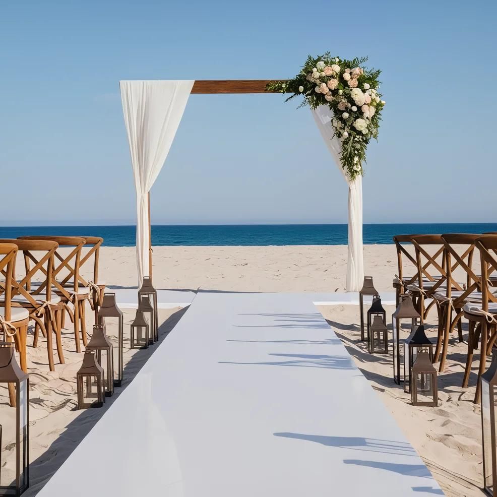 A beach wedding setup with an aisle leading to an archway decorated with white drapes and floral arrangements. Wooden chairs and lanterns line the sides of the aisle.