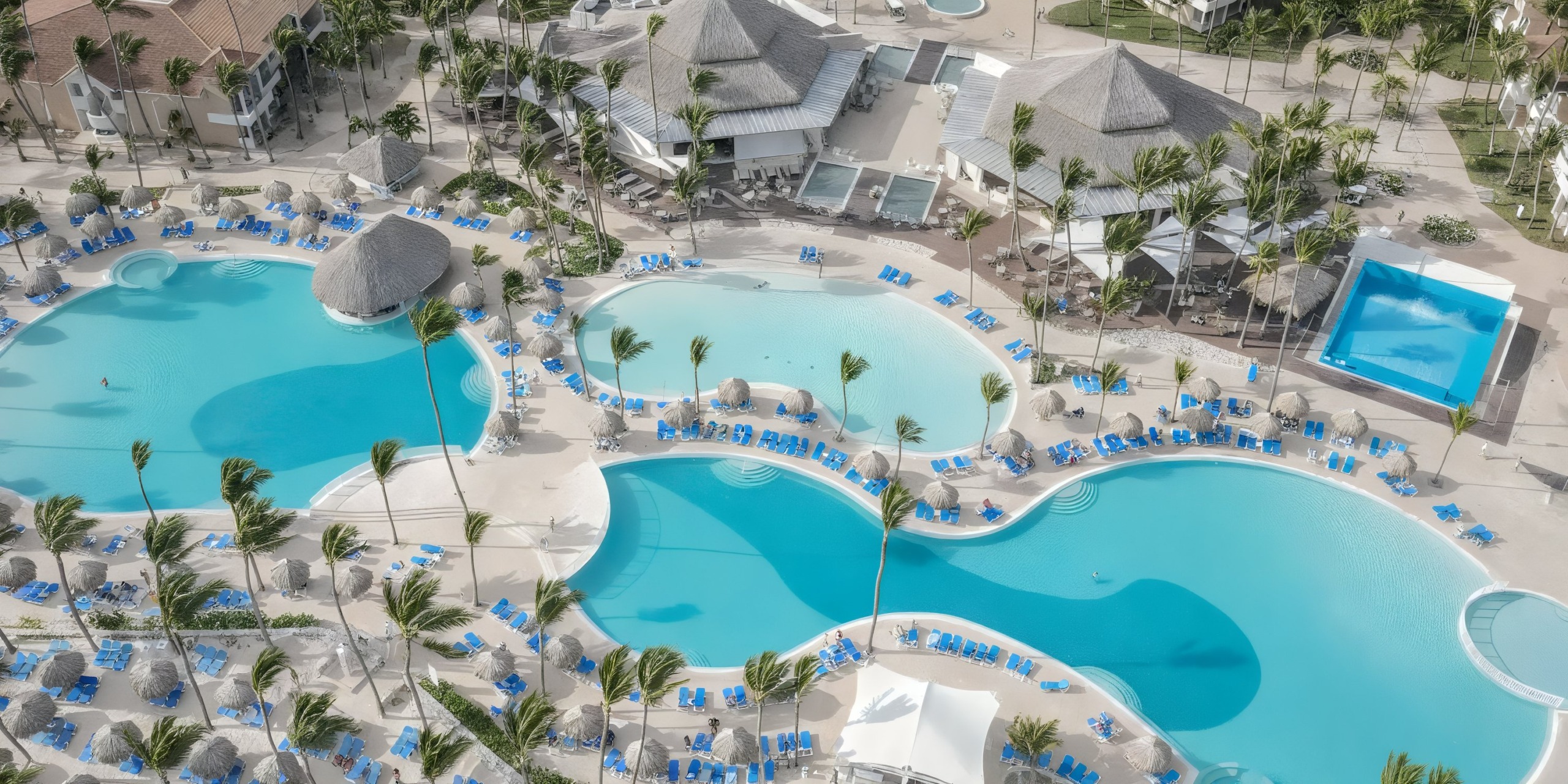 Aerial view of Bahia Principe Luxury Ambar resort, featuring lagoon-shaped pools, palm trees, blue lounge chairs, and nearby thatched-roof structures.