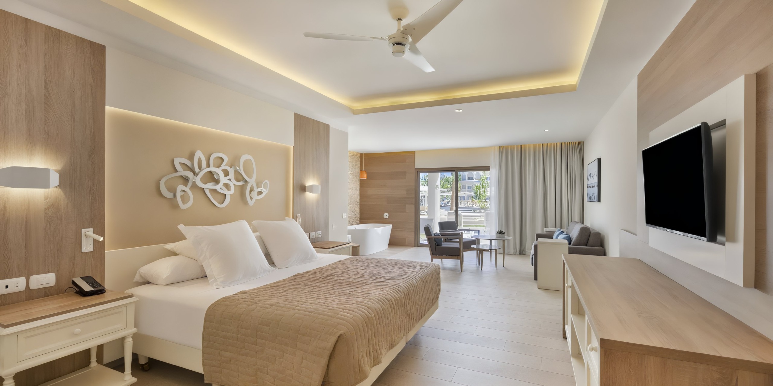 Experience a spacious room at Bahia Principe Luxury Ambar featuring a king-size bed, modern decor, wall-mounted TV, seating area, ceiling fan, and a large window opening to a balcony with stunning views.