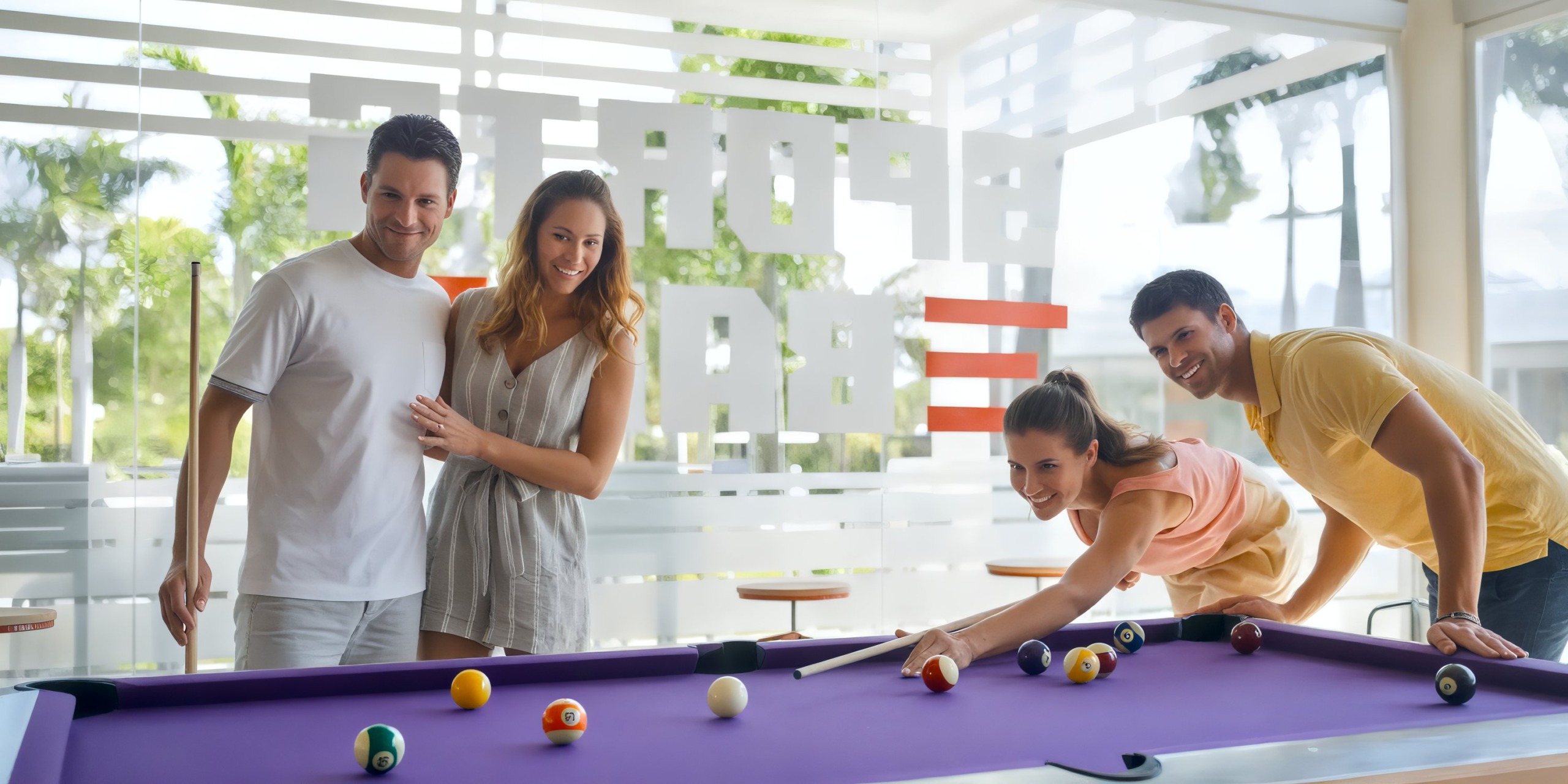 Four people play pool on a purple table at Bahia Principe Luxury Ambar in a bright, casual setting with large windows.