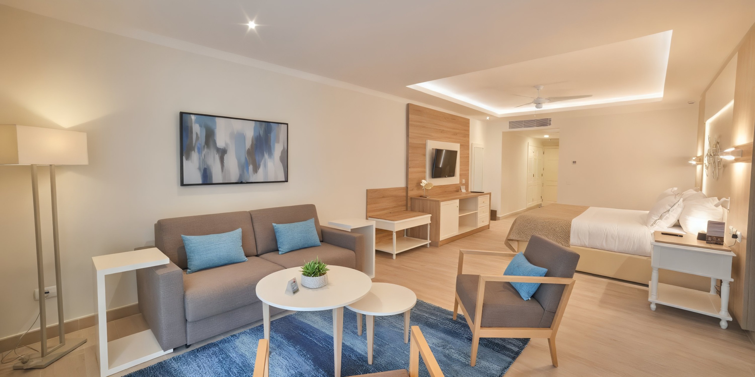 The Bahia Principe Luxury Ambar hotel room features a modern design with a sofa, armchairs, and a bed. The decor includes wall art, a floor lamp, and a TV, with neutral colors highlighted by blue accents.